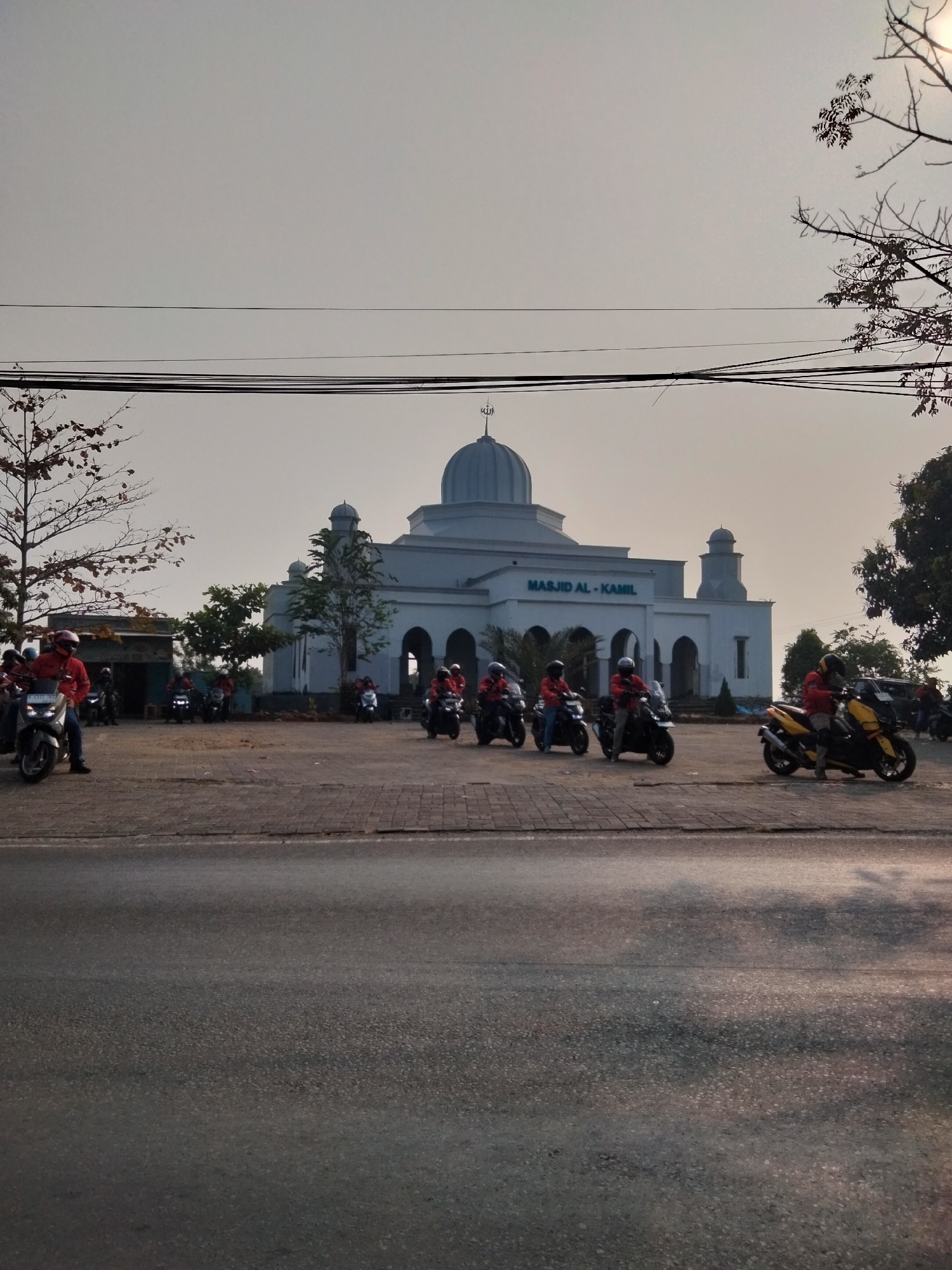A group of motorcyclists are parked in front of the Masjid Al-Kamil in Cariu, Indonesia. The mosque is a white building with a dome and minarets, and the name of the mosque is visible in green lettering above the entrance. The sky is overcast, and the sun is setting, casting a warm glow on the scene. The motorcyclists are wearing helmets and red jackets, and they appear to be taking a break from their ride. The image also shows trees and power lines in the background. The road in the foreground is asphalt, and there is a paved walkway in front of the mosque.