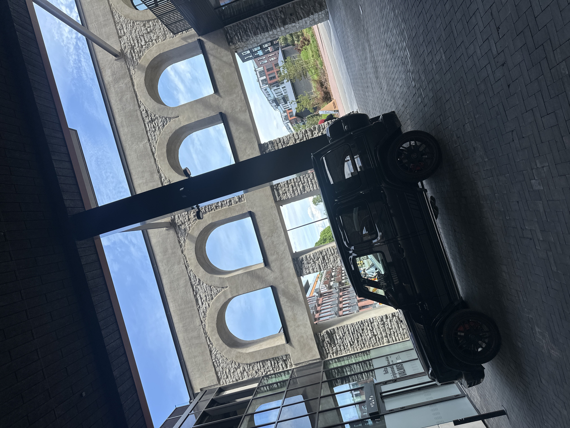 The image depicts a black Mercedes-Benz G-Wagen parked on a patterned brick walkway, likely in a modern urban setting. The vehicle is positioned at an angle, facing towards the right of the frame, with its rear tire and spare tire cover visible. The background features a contemporary building with arched windows that reveal a blue sky with scattered clouds. Adjacent to this structure is a glass-fronted building with reflections of the sky and surroundings. Further in the distance, across a street, are several multi-story residential buildings with a mix of architectural styles. This scene is consistent with the architecture found in the Volta neighborhood of Tallinn, Estonia, known for its redevelopment of former industrial areas into modern residential and commercial spaces. No people are clearly visible interacting with the vehicle or within the immediate vicinity. The lighting suggests it is daytime, with shadows indicating the sun is overhead.