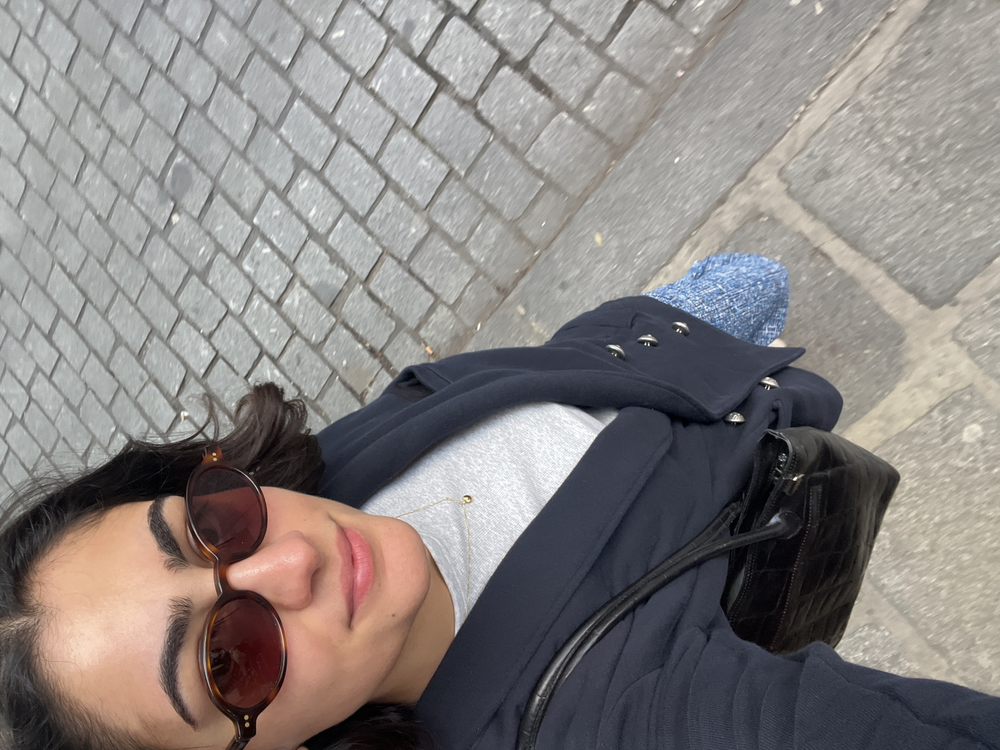 A woman is shown in a selfie-style, tilted head-and-shoulders shot from above. She is wearing brown round sunglasses. Her hair is dark. She wears a gray top, a dark blue coat, and a gold necklace. She carries a small black leather bag with a shoulder strap. The background shows a cobblestone street and part of a concrete sidewalk. Based on the distinctive cobblestone pattern, this image was likely taken in Paris, France.