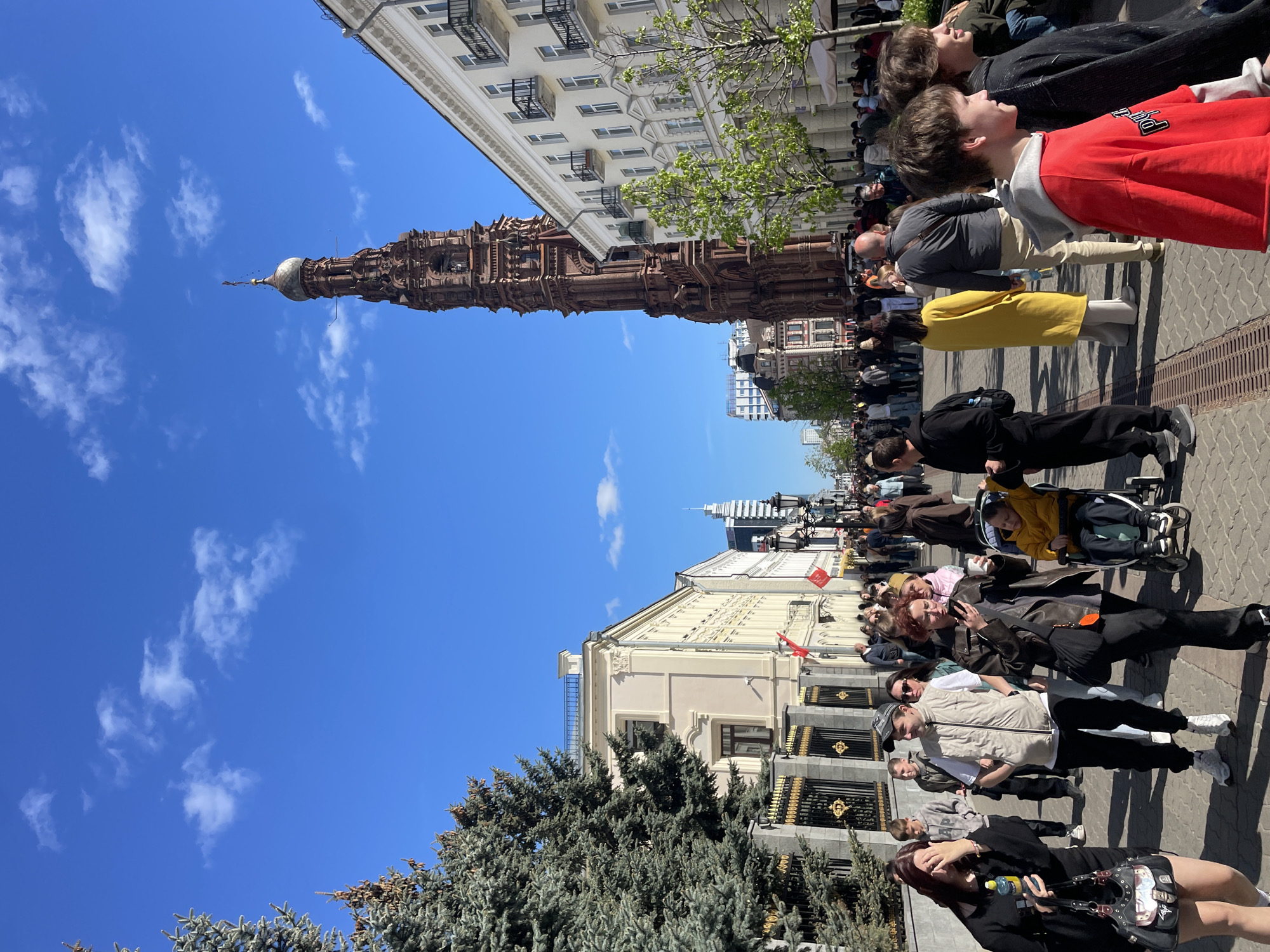 The image depicts a daytime outdoor scene in Kazan, Russia, showcasing the Soyembika Tower against a bright blue sky with scattered clouds. The tower, a tall, brown brick structure with a golden dome, is prominently featured on the left side of the image.
A crowd of people, varying in age and attire, is visible on the right side of the image, suggesting a public space or gathering. Trees with blueish foliage are in the foreground. Buildings line the street with a mix of architectural styles. A red flag is visible on one building. Some individuals are looking upwards towards the tower.