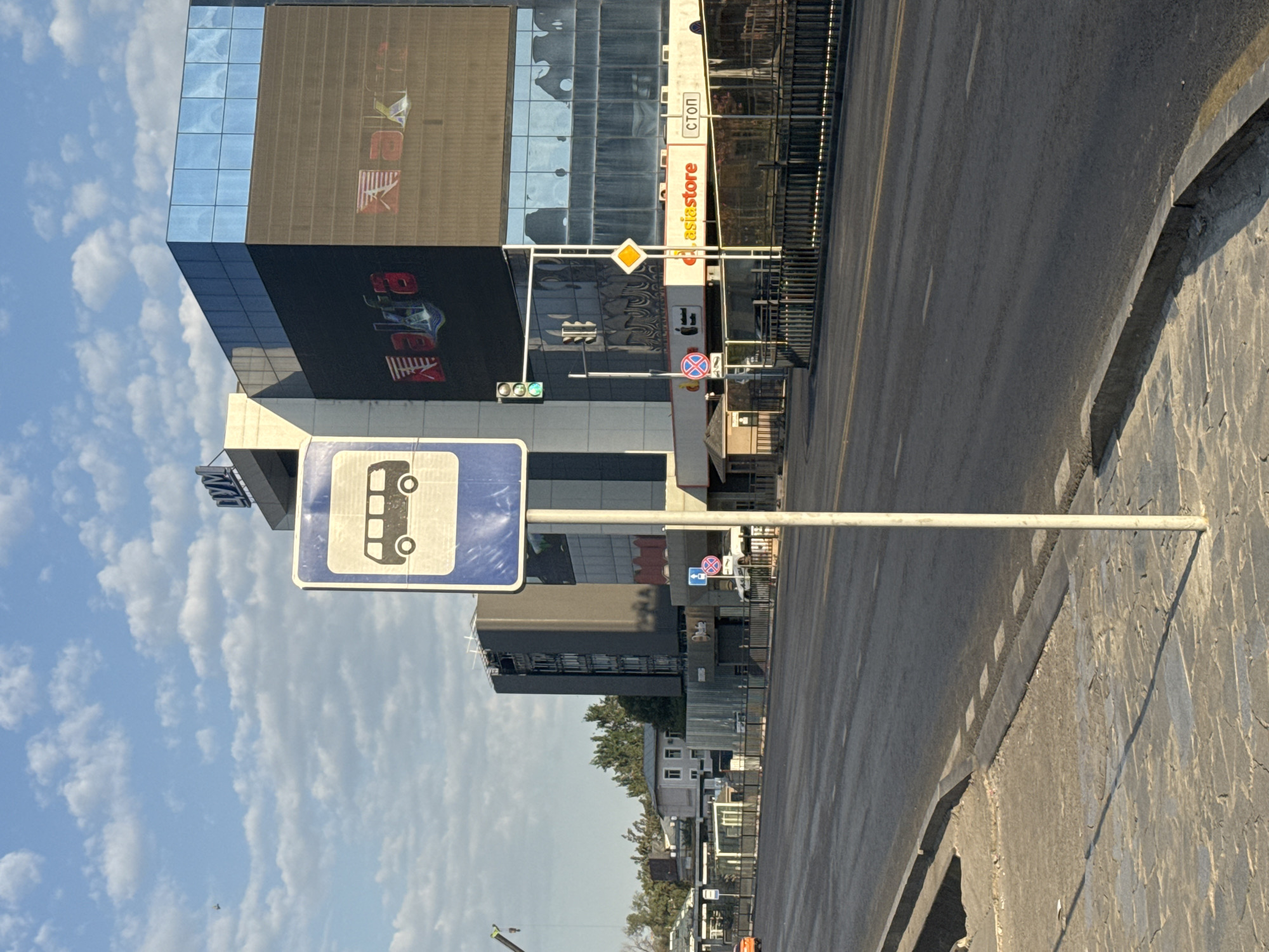 A bus stop sign stands on a sidewalk next to a road in Bishkek, Kyrgyzstan. The sign is blue and white with a pictogram of a bus. Behind the sign, a large, modern building with a glass facade dominates the background. The building has "nalfa" and "Asia Store" signage visible. A traffic light is also present near the building. The road is asphalt with lane markings. The sky is blue with scattered clouds. The scene appears to be a typical urban streetscape.