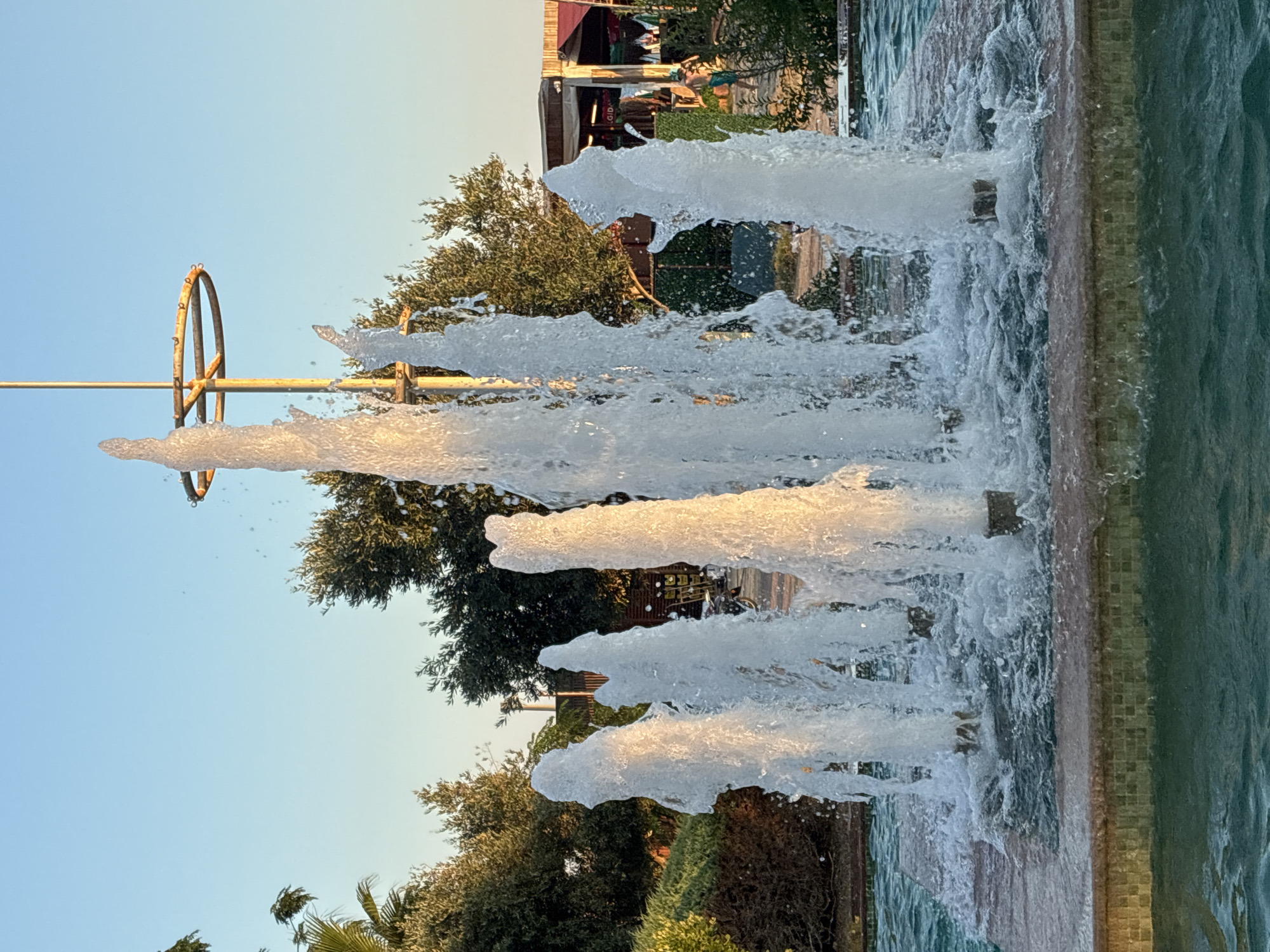 The image shows a fountain with multiple water jets in Alanya, Turkey. The fountain is in a shallow basin with tiled edges. In the background, there are trees, buildings, and people. The sky is clear and blue. The water jets are illuminated by the sun, creating a shimmering effect. One jet is significantly taller than the others and appears to be capped with a metal ring. The scene appears to be a public space, possibly a park or a plaza, in Alanya.