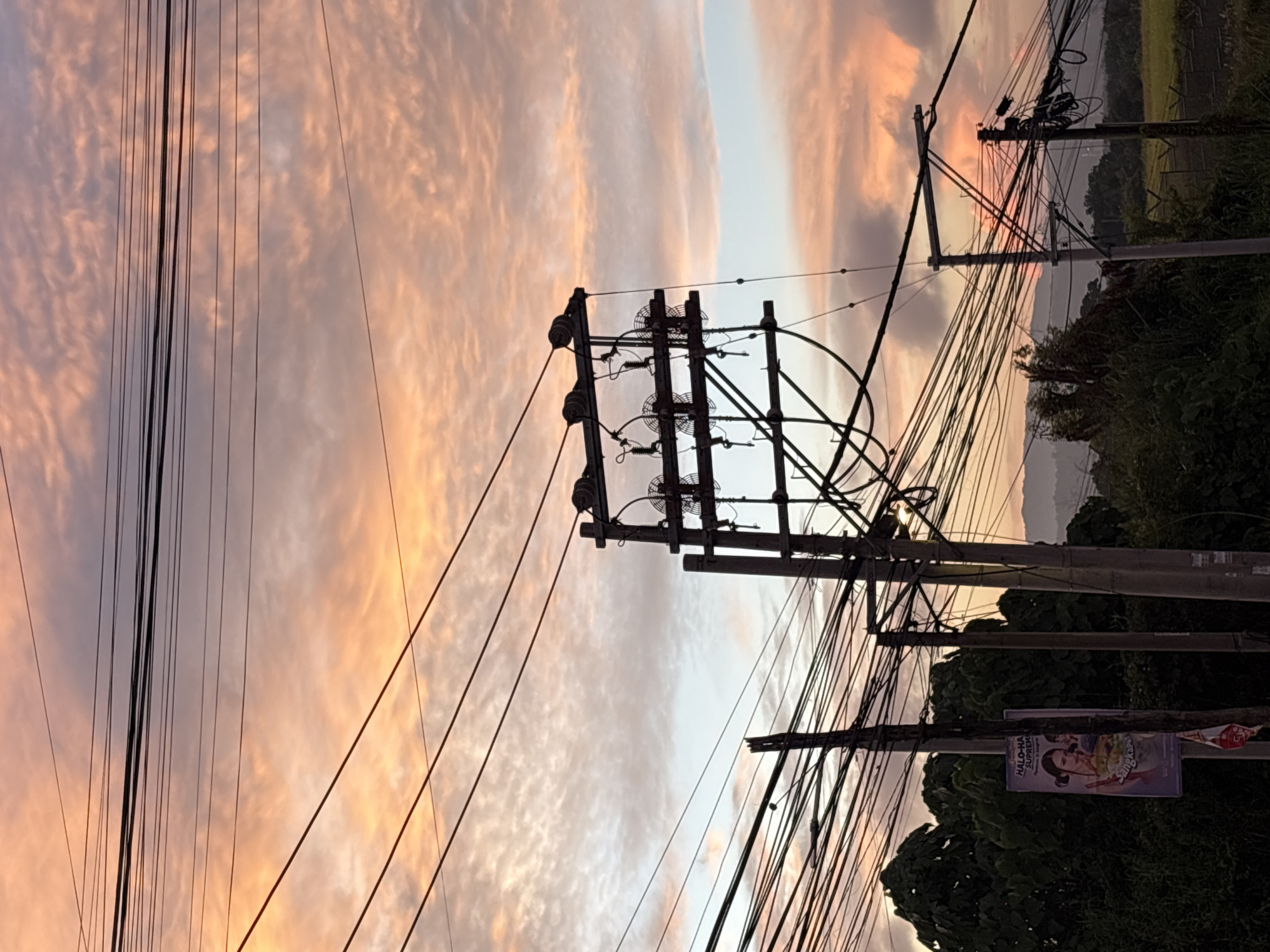 The image depicts a street scene in Davao City, Philippines, characterized by a dramatic sunset sky filled with streaks of pink, orange, and gray clouds. Prominent electrical poles and a dense network of overhead wires dominate the foreground, crisscrossing the frame and partially obscuring the sky. On the left side, a poster advertising "Halo-Halo Supreme" is attached to a pole, featuring an image of a woman holding a dessert. Lush green trees and foliage are visible behind the poles, and in the distance, rolling hills and mountains are faintly silhouetted against the horizon. The overall impression is a blend of urban infrastructure and natural beauty, captured during the twilight hours.