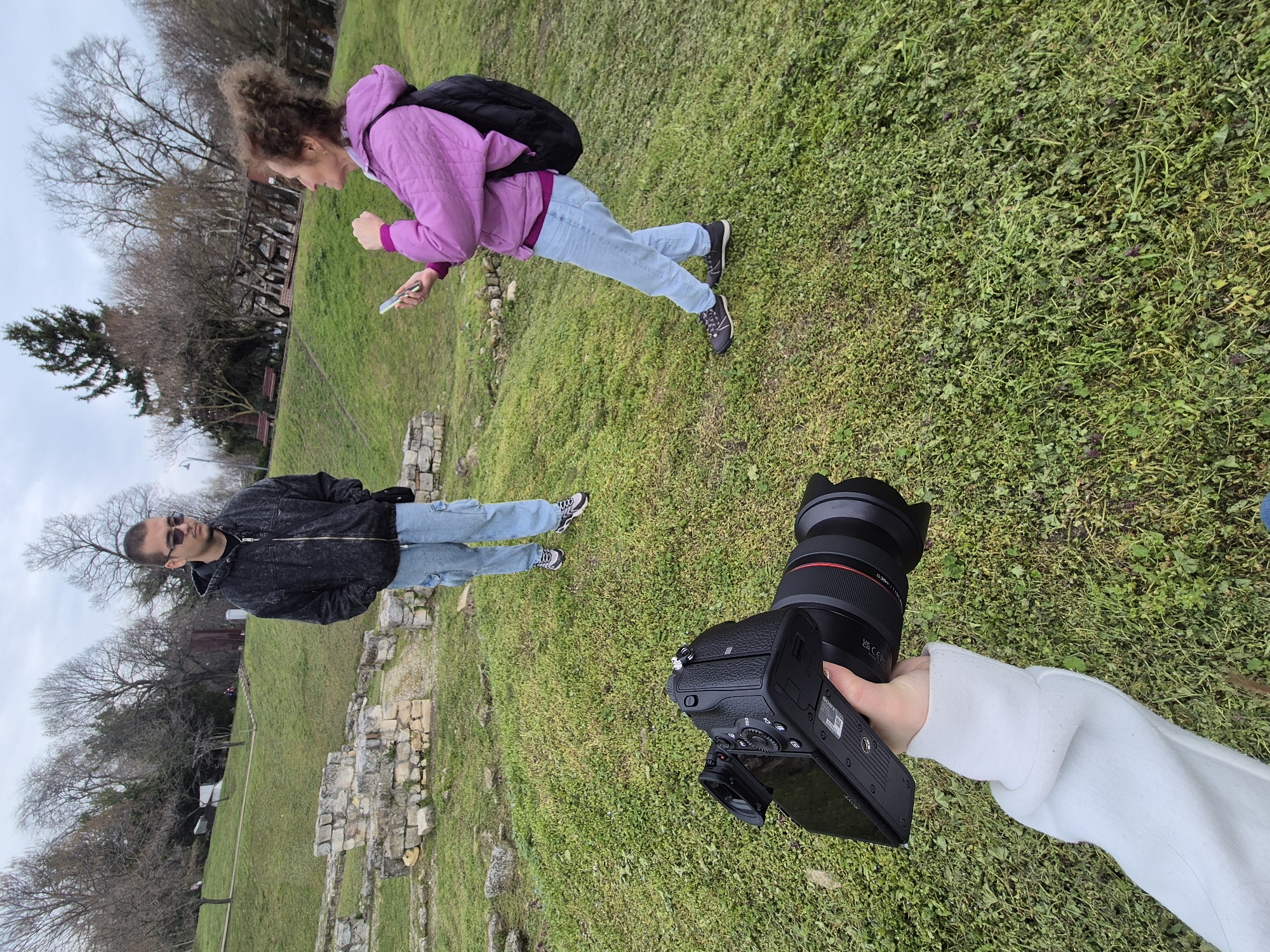 The image shows a park with Roman ruins in Silistra, Bulgaria. Two people are in the image. The first is a person with light curly hair, wearing a pink jacket, light blue jeans, and a black backpack. They are looking at a phone in their hand. The second person is wearing a black jacket, light blue jeans, and sunglasses. They are standing near the ruins. In the foreground, a person holds a Sony digital camera. The area is covered in green grass. Bare trees are visible in the background. The sky is overcast.