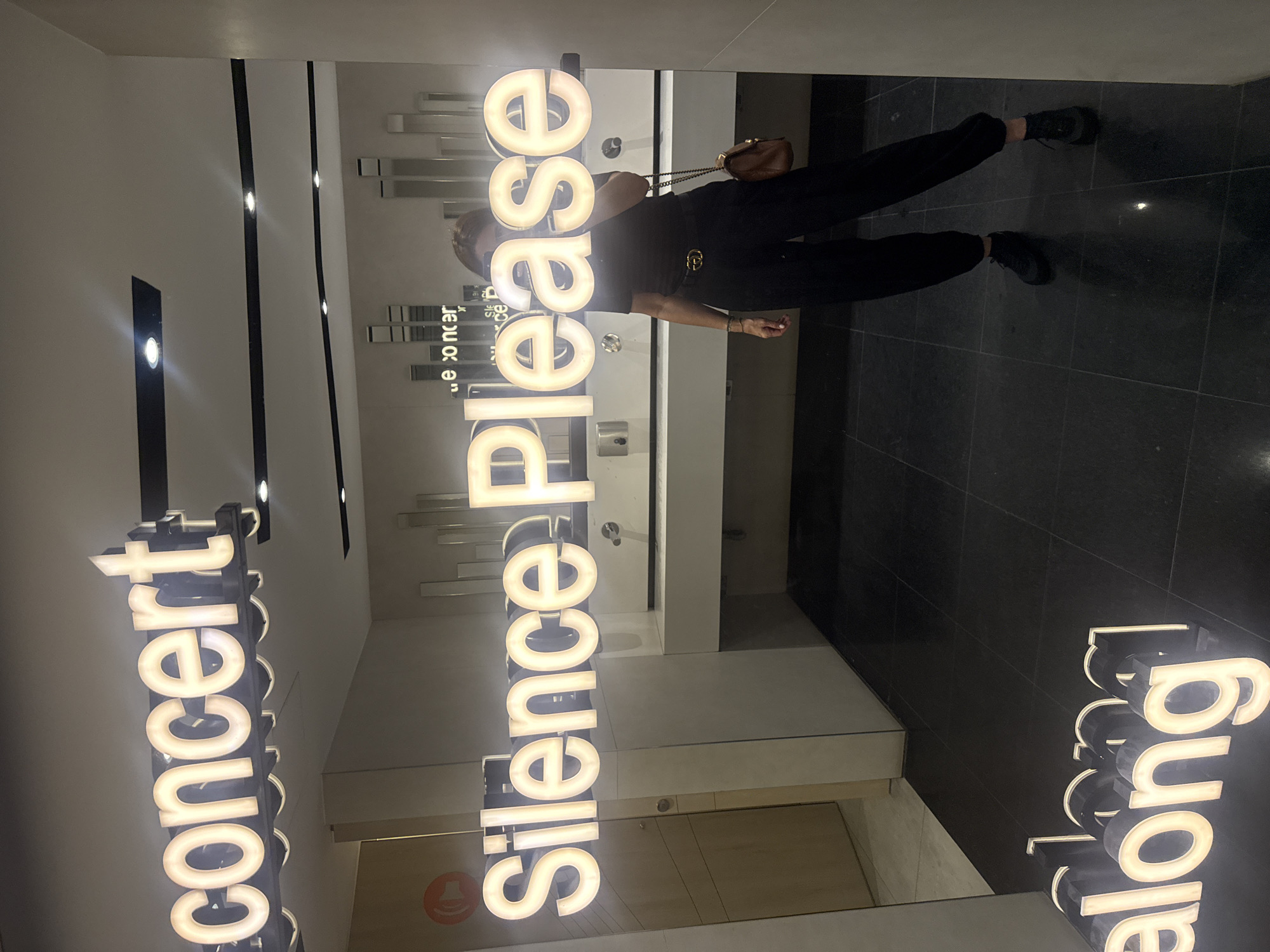 A woman in black clothing is reflected in a mirror in a building in Vila Nova de Gaia, Portugal. The mirror reflects her torso, legs, and part of her face. She has a brown purse and is wearing black sneakers. The words "concert," "silence Please," and "along" are displayed in illuminated vertical signs in the mirror's reflection. The interior is modern with light-colored walls and dark flooring. There are modern, rectangular decorative mirrors on the wall.
