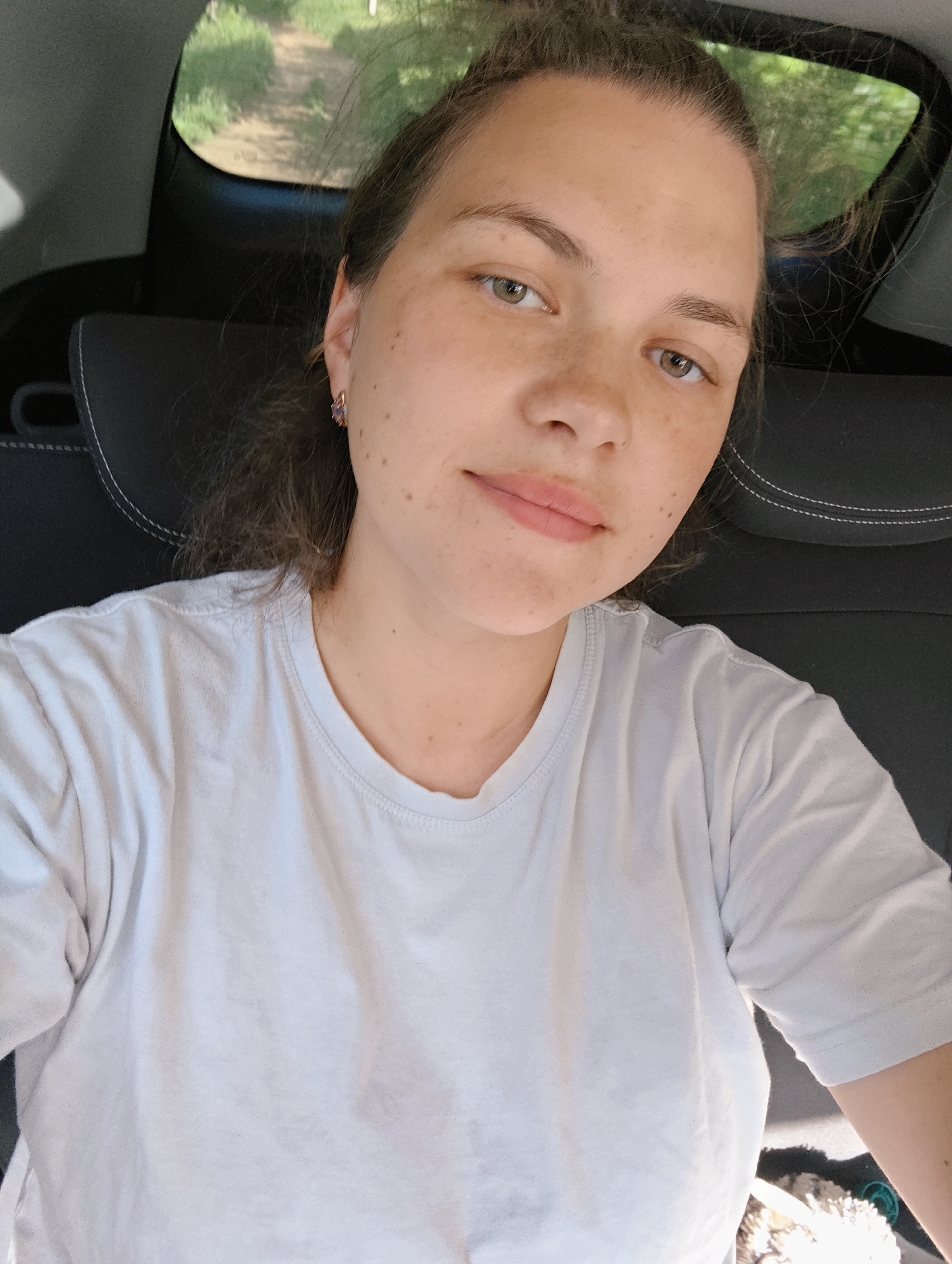 A fair-skinned woman with light brown hair pulled back is in the backseat of a car. She wears a plain white t-shirt and has a small earring in her left ear. The car seat is dark with light-colored stitching. Through the car window, a dirt road and greenery are visible. The location is Zhigalki, Russia.