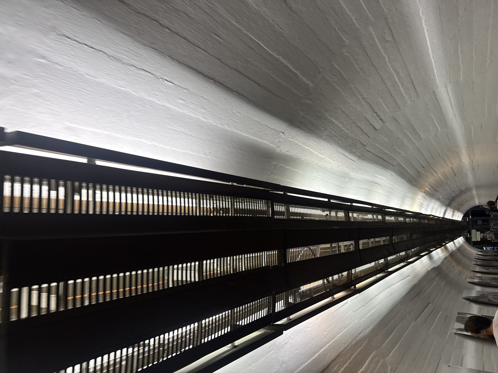 The image shows a perspective shot looking up into a tunnel or underpass. The ceiling is a dark, slatted structure with lighting visible through the slats, creating a striped effect. The walls of the tunnel are rough, white-painted brickwork, curving upwards and inwards. There are faint shadows cast on the wall from the slatted ceiling. In the distance, there appears to be more people and possibly an exit or different section of the tunnel. Some framed artworks are visible on the left wall of the tunnel. The image does not provide any specific details or landmarks that would allow for identification of the city of Yalta, Ukraine.