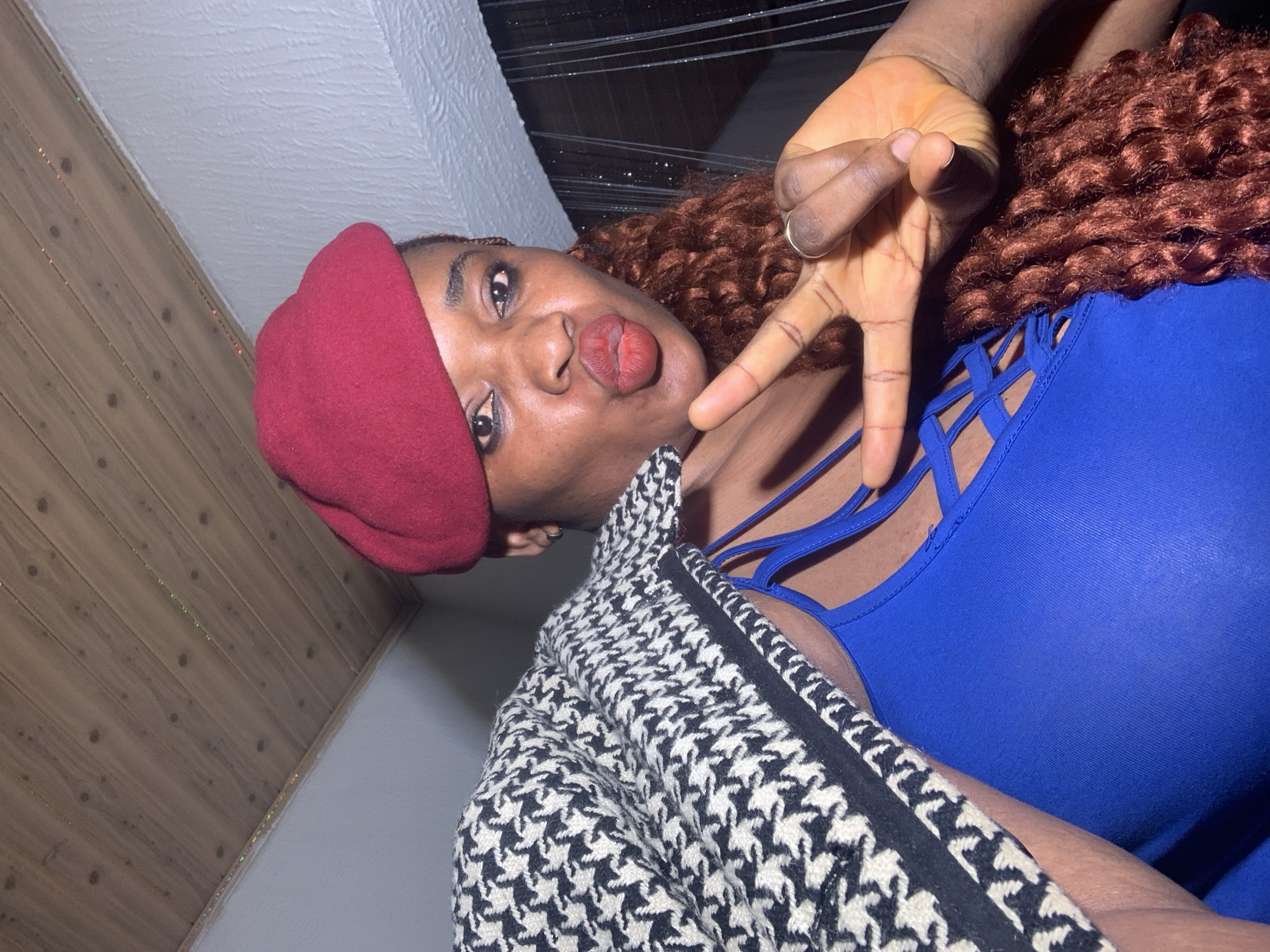 A young woman is captured in a close-up, angled shot, likely a selfie, in an indoor setting. She has dark skin, dark eyes, and a mass of curly, reddish-brown hair on her right side. She is wearing a deep red beret on her head, a vibrant blue top featuring intricate criss-cross strap detailing across the chest, and a black and white houndstooth patterned jacket draped over her shoulders. Her lips are puckered into a playful expression, and her gaze is directed towards the camera. Her right hand is raised, making a "peace" or "V" sign, with a gold-toned ring visible on her ring finger.
The background reveals a room with an angled design. To the left, there is light brown wooden paneling, possibly on a wall or ceiling, featuring subtle reflective specks. To the right, a white, textured surface, either a wall or ceiling, is visible. The lighting is artificial and appears to be ambient, typical of an indoor environment. The woman's pose and facial expression convey a sense of playfulness and confidence. There is no visible text in the image. The location context for this image is Barakin Akawo, Nigeria.