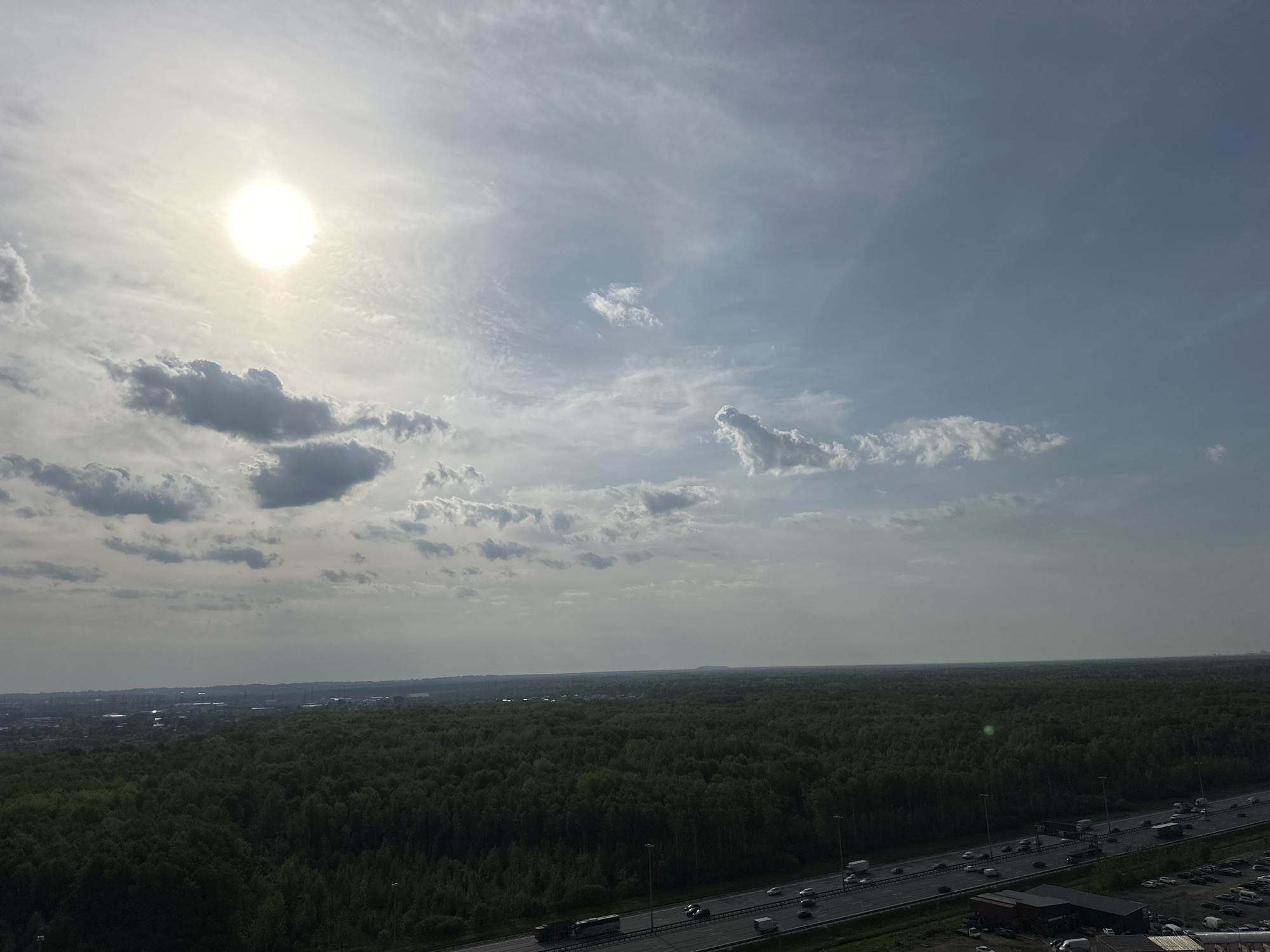 The image shows an aerial view of a highway and a large wooded area on what appears to be a partly cloudy day. The sun is visible in the upper left corner, partially obscured by clouds. A multilane highway runs across the lower right portion of the image with cars and trucks traveling in both directions. To the left of the highway, there appears to be a large expanse of dense forest, possibly extending to the horizon. In the distance, there are indications of a city skyline, suggesting this location is on the outskirts or edge of a metropolitan area. The location in the city of Unknown, Unknown, cannot be discerned from the image.