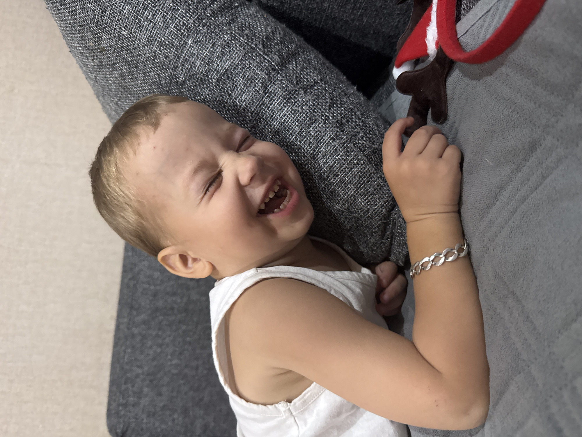 A young Caucasian boy with blond hair and blue eyes is lying on his side on a gray sofa, laughing. He is wearing a white tank top and a silver bracelet on his left wrist. His eyes are closed, and his mouth is open, showing his teeth. Next to him is a pair of reindeer antlers with a red and white Santa hat attached to them. The boy is positioned in the foreground, with the sofa and a wall visible in the background. The scene appears to be indoors, possibly in a home. The overall impression is one of joy and playfulness.