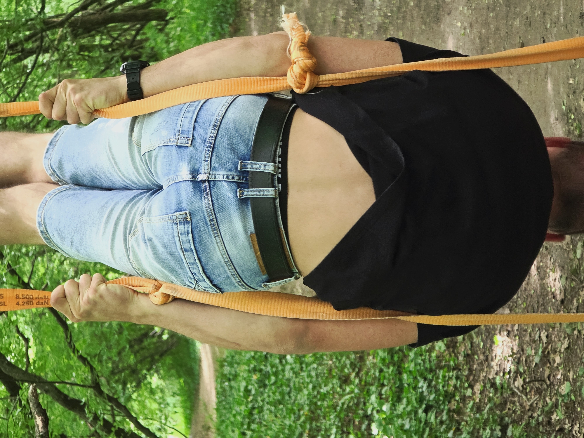 A man, photographed from above in an outdoor setting, is straddling a slackline made of a flat, orange strap. He wears blue jean shorts and a black t-shirt. The slackline is taut, held at both ends by his hands. He also wears a black watch on his left wrist. The background reveals a wooded area with trees and a visible path. Based on prior information, it can be inferred the location is Shumen, Bulgaria.