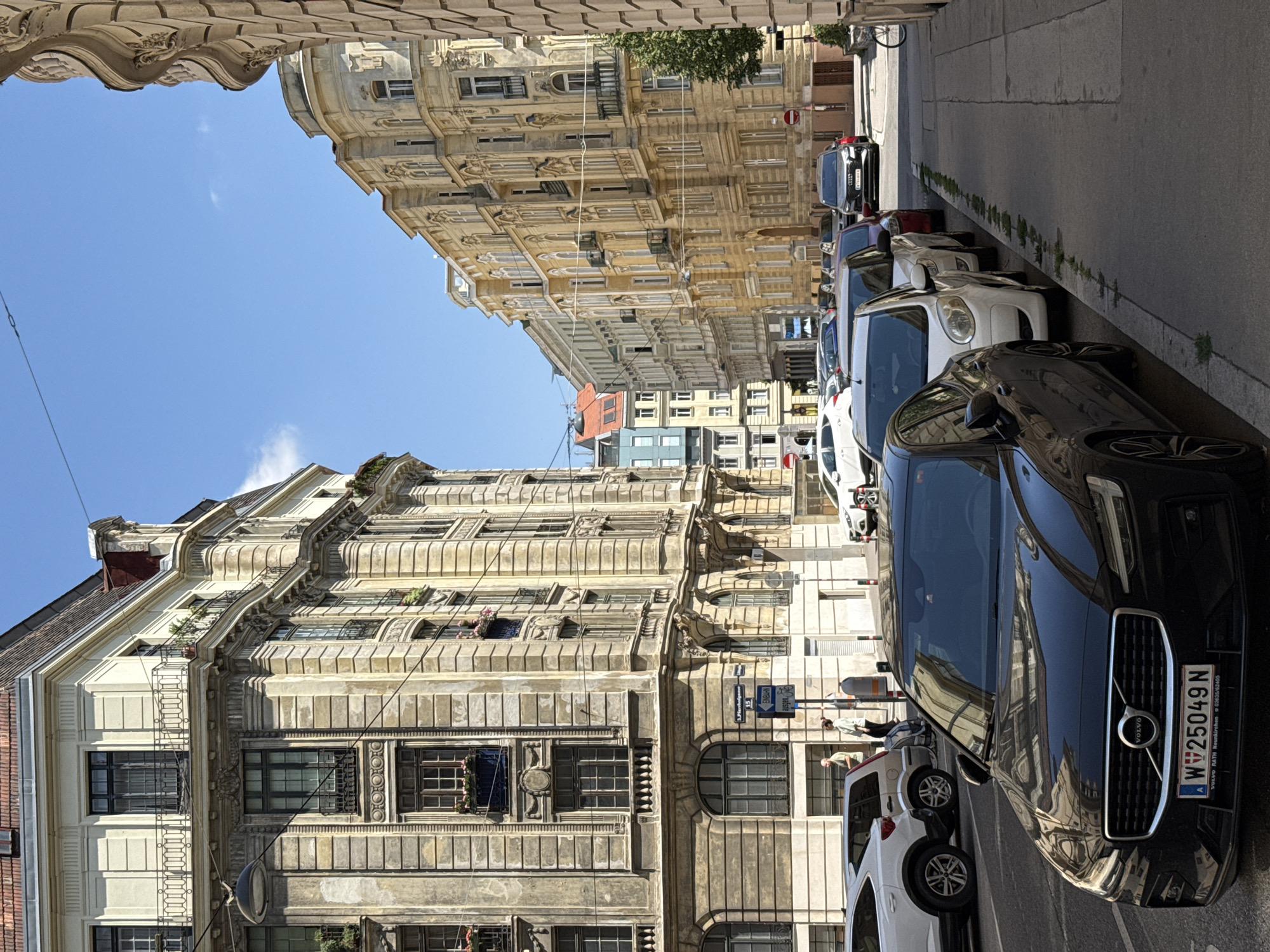 This image captures a street scene in Vienna, Austria, likely on a bright, sunny day given the clear blue sky and sharp shadows. The foreground is dominated by the front of a black Volvo sedan, identifiable by its license plate, which reads "W V25049 N". Behind it, several other vehicles are parked along the street, including a white Citroen and a dark red car.
The street is lined with ornate, multi-story buildings characteristic of Viennese architecture. The building on the left, with its detailed facade, appears to be older, featuring a classical style with balconies and numerous windows. The buildings across the street are also grand, painted in shades of yellow and cream, with intricate detailing around the windows and rooflines.
A few pedestrians are visible in the mid-ground, near a street sign. One person, wearing a light-colored shirt and dark pants, appears to be walking. Another individual in a light green top and dark pants stands near a street sign. The street itself is paved, with a sidewalk running alongside the buildings. A lamppost is visible on the sidewalk. Further down the street, more buildings and parked cars can be seen, extending into the distance. The overall impression is of a well-maintained, historic urban environment.
