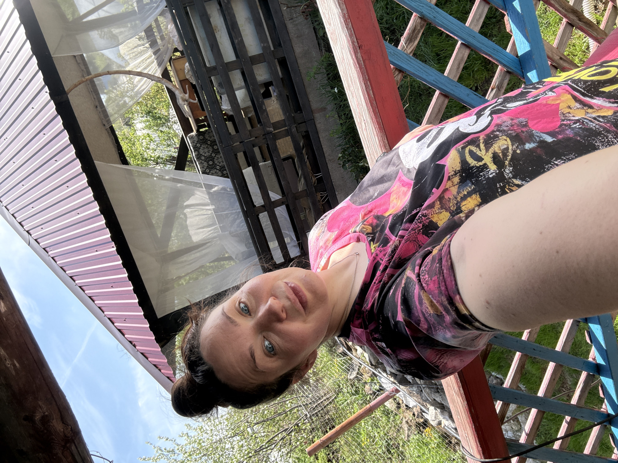 A medium-shot selfie depicts a woman with fair skin, blue eyes, and dark hair pulled back into a bun. She is wearing a t-shirt with a black background and colorful patterns in pink, yellow, and white. The woman appears to be situated outdoors, near a red and blue painted wooden structure resembling a fence or trellis, with green grass visible in the background. Over her head, there is a roof constructed with metal sheets and a transparent covering, possibly part of a gazebo or similar outdoor structure. A wooden lattice or pallet is visible near the roof. The photo was taken in the city of Chaikovsky, Russia.