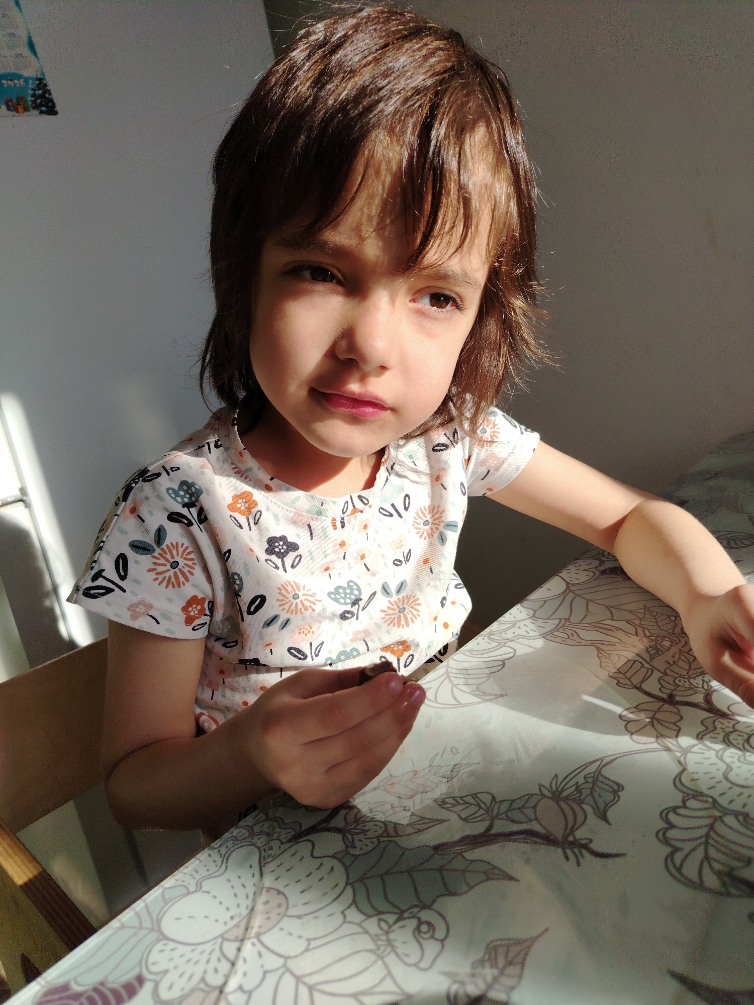 A young girl with brown hair and eyes sits at a table. She is wearing a white shirt with a floral pattern. She holds a small, dark object in her hand. The table has a patterned tablecloth with a light background. The background is a white wall, with a partially visible object hanging on the wall. A wooden chair is visible on the left side of the frame. The scene is lit with natural light, casting shadows. There is no information in the image to indicate the location in the city of Noviy, Russia.