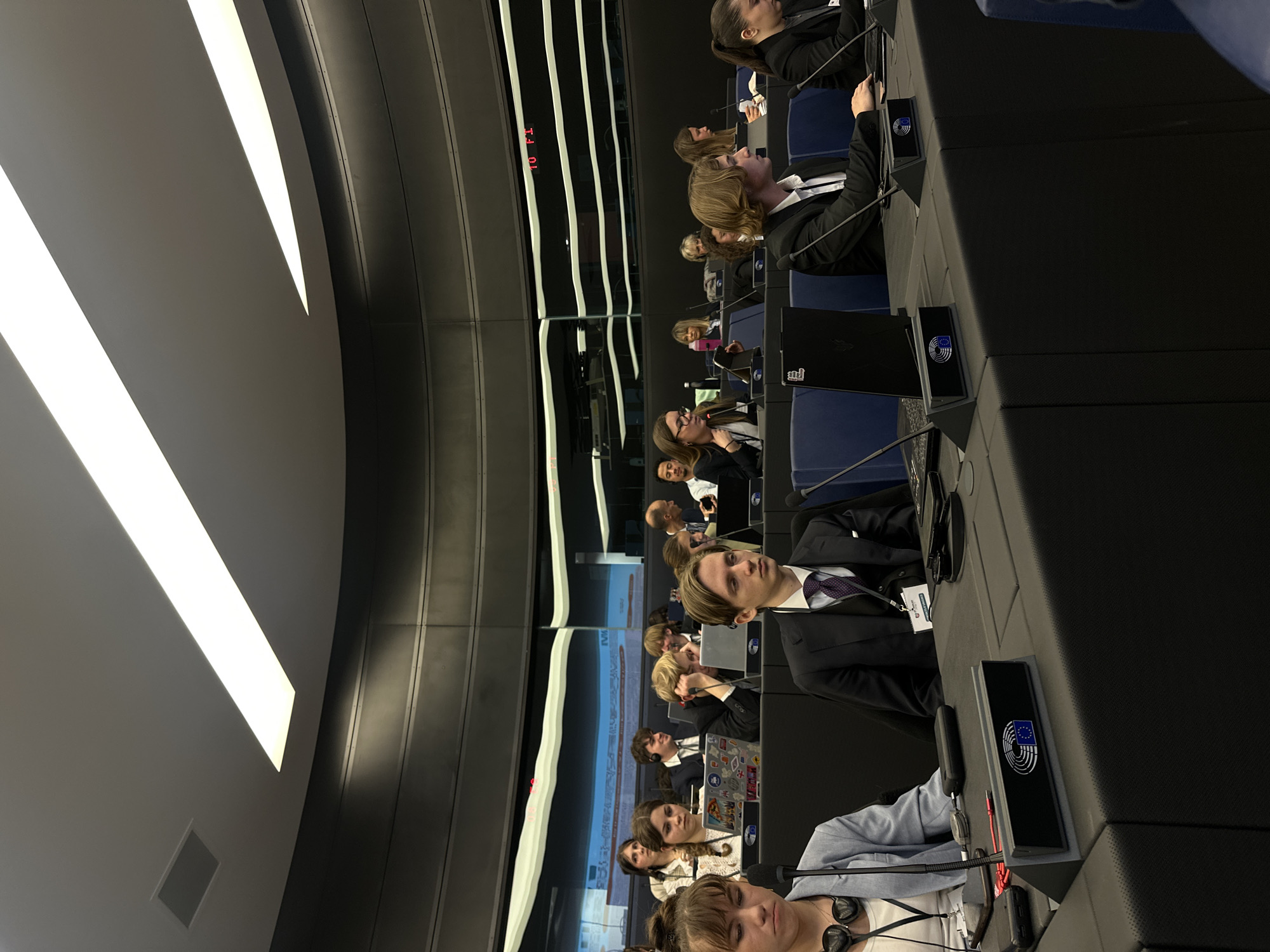 The image shows the interior of the European Parliament in Strasbourg, France, with rows of seated individuals, some wearing suits, at desks equipped with monitors displaying the European Union emblem. The setting appears to be a plenary session or similar assembly, as indicated by the organized seating and the devices present on the desks. The room is illuminated by modern, linear lighting fixtures, and a display panel with red numbers is visible on the wall.