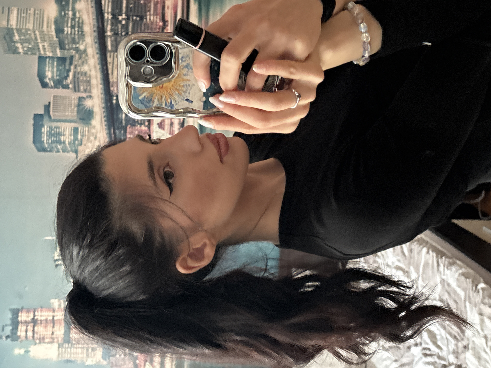 A woman with dark hair pulled back in a ponytail is holding a silver smartphone with a floral-designed case in her left hand and a black object, possibly lipstick or a compact, in her right. She's wearing a black long-sleeved shirt, a beaded bracelet on her left wrist, and a ring on her right hand. Her nails are painted with a white French tip. The background appears to be a cityscape. There is no evidence to suggest the picture was taken in Sofia, Bulgaria.