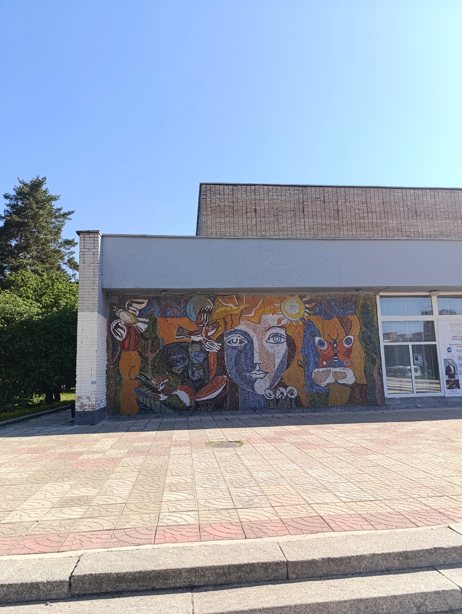 The image shows the facade of a building in Chernogolovka, Russia, featuring a large mosaic mural. The mural is composed of many small colored tiles and depicts a stylized human face surrounded by various natural elements such as birds, foxes, and a sun. The building itself appears to be made of brick and has a modern architectural style. The foreground shows a paved area with patterned tiles, and steps leading up to the building's entrance. The sky is blue, and a tree is visible to the left of the building.
