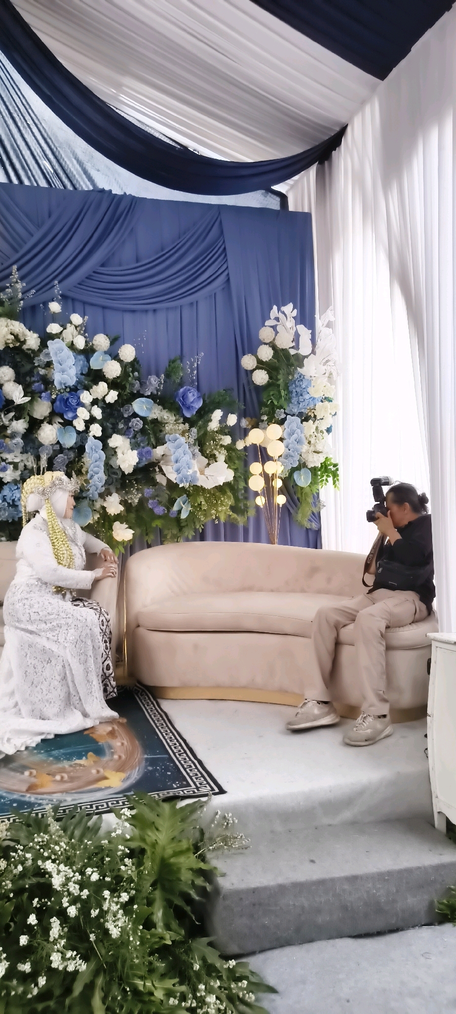 In South Tangerang, Indonesia, a wedding ceremony is taking place, captured by a photographer. A bride, dressed in a white lace gown and traditional headwear, is seated on a plush, light-colored sofa. Her attire features intricate embellishments, and she wears a veil. Beside her, on the same sofa, is a dark, patterned textile. In front of the bride and sofa, a dark rug with an intricate design is laid out.
To the right of the bride, a photographer, positioned on the floor, is actively taking pictures. The photographer is wearing a black top and tan pants, with a camera bag slung across their shoulder. They are focused on the bride, holding a professional camera with a flash unit attached.
The backdrop consists of voluminous blue drapes that create a layered effect. Adorning the blue drapes are abundant floral arrangements, featuring a mix of white and various shades of blue flowers, interspersed with greenery. To the right, white sheer curtains hang, adding a soft, diffused light to the scene. In the mid-ground, near the floral arrangements, a cluster of spherical, illuminated lamps on thin rods adds a decorative element. The floor is a pale, reflective surface, with a hint of green foliage visible in the lower foreground.