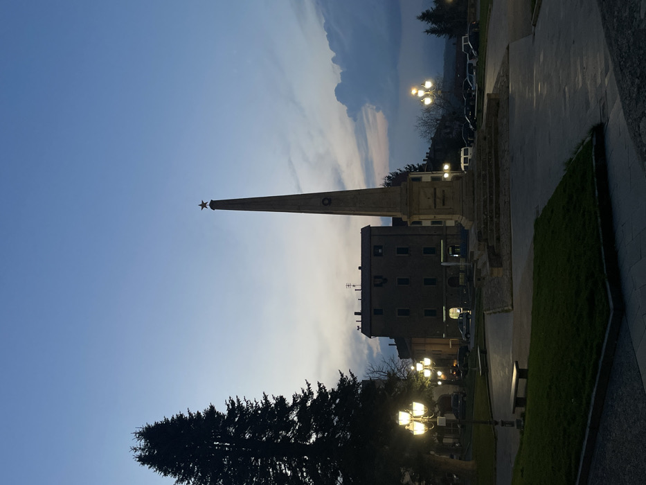 The image displays a public square in Castel del Piano, Italy, at dusk. A tall, light-colored stone obelisk, crowned with a five-pointed star and featuring a wreath emblem on its shaft, occupies the center, resting on a multi-tiered base. To the left of the obelisk, a large, dark evergreen tree stands prominently. The foreground features manicured dark green grass areas bordered by light-colored paved walkways. A dark park bench is visible on a walkway to the left, alongside a four-lamp street light emitting yellow illumination. In the mid-ground, behind the obelisk, a multi-story building with dark facades and illuminated ground-floor windows is present. To the right of the obelisk, a three-lamp street light illuminates a parking area containing multiple parked vehicles, including cars and a white van. The sky transitions from deep blue overhead to lighter hues near the horizon, with some indistinct clouds visible on the right. No individuals are present in the scene.
