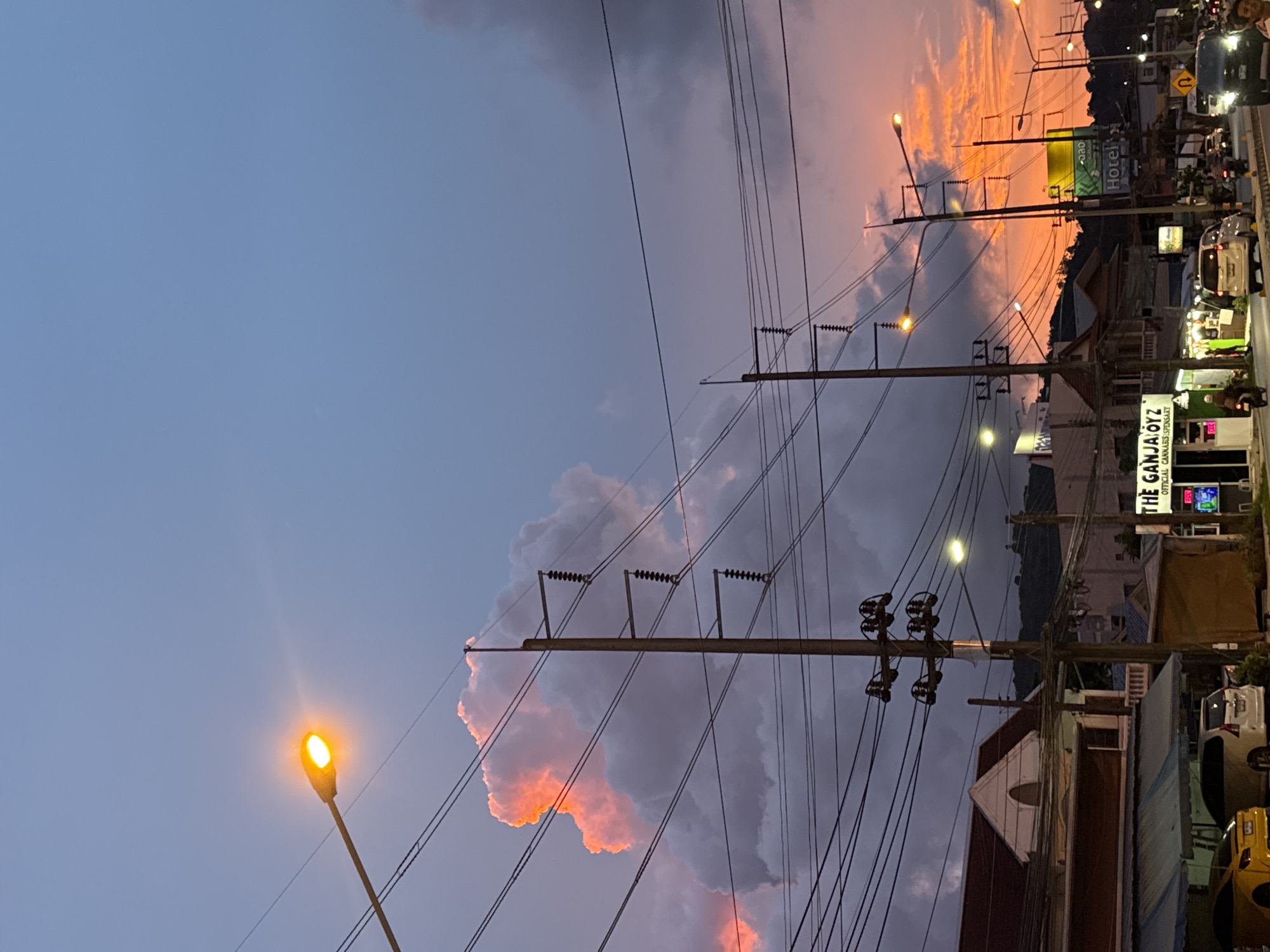 The image depicts a street scene at dusk in an urban environment. The sky is a gradient of deep blue to vibrant orange, with illuminated clouds contributing to the dramatic sunset. Numerous utility poles with tangled wires crisscross the frame, interrupting the view of the sky and the buildings below.
On the left side of the street, a building with a pitched roof and a dormer window is visible, with awnings extending over a storefront. Next to it, another building houses a business with neon signage that reads "THE GANJA OYZ OFFICIAL CANNABIS DISPENSARY." A yellow car is parked in front of one of the establishments, and a white van is also visible. Several motorcycles are present on the street, with one rider in motion.
The right side of the street features more buildings, including one with a sign that says "Hotel." Streetlights are illuminated, casting a warm glow on the scene. Traffic is present on the road, with various vehicles, including cars and motorcycles, moving along. The overall atmosphere is that of a bustling street during the transition from day to night, characterized by the vibrant colors of the sunset and the man-made infrastructure of the city.