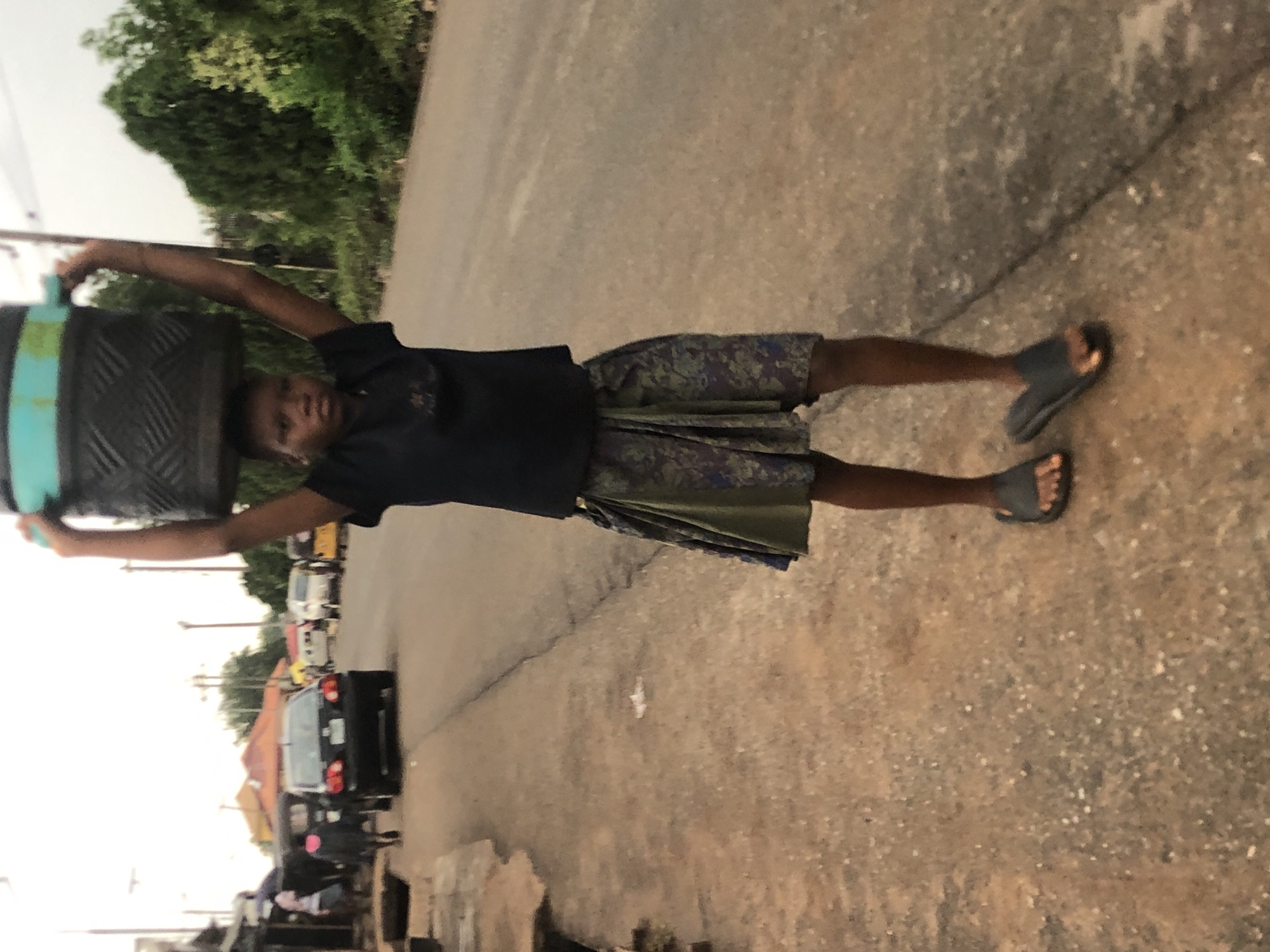 A full-body shot captures a young person balancing a large, dark, cylindrical object on their head while standing on a paved road in Nsugpe, Nigeria. The image is oriented vertically, with the person upright.
The subject, appearing to be a young boy with dark skin, looks directly at the camera with a neutral expression. He wears a dark, short-sleeved shirt and patterned shorts or a wrap-around skirt in shades of green, blue, and brown. His arms are raised to steady the heavy load, which appears to be a tire or a drum, marked with a light blue or teal band. He is wearing dark sandals on his feet.
The setting is an outdoor street or road with a light-colored, cracked paved surface. To the person's right, there is a dirt shoulder. The background features lush green trees and foliage. Further down the road, several vehicles, including cars and vans, are visible, suggesting a busy thoroughfare or town area. Power lines and utility poles are also present in the distance.
The activity depicted is the act of carrying a heavy load on one's head, a traditional method of transport. The lighting suggests it is daytime, possibly an overcast afternoon, given the soft, diffused light and lack of harsh shadows. There is no visible text in the image.