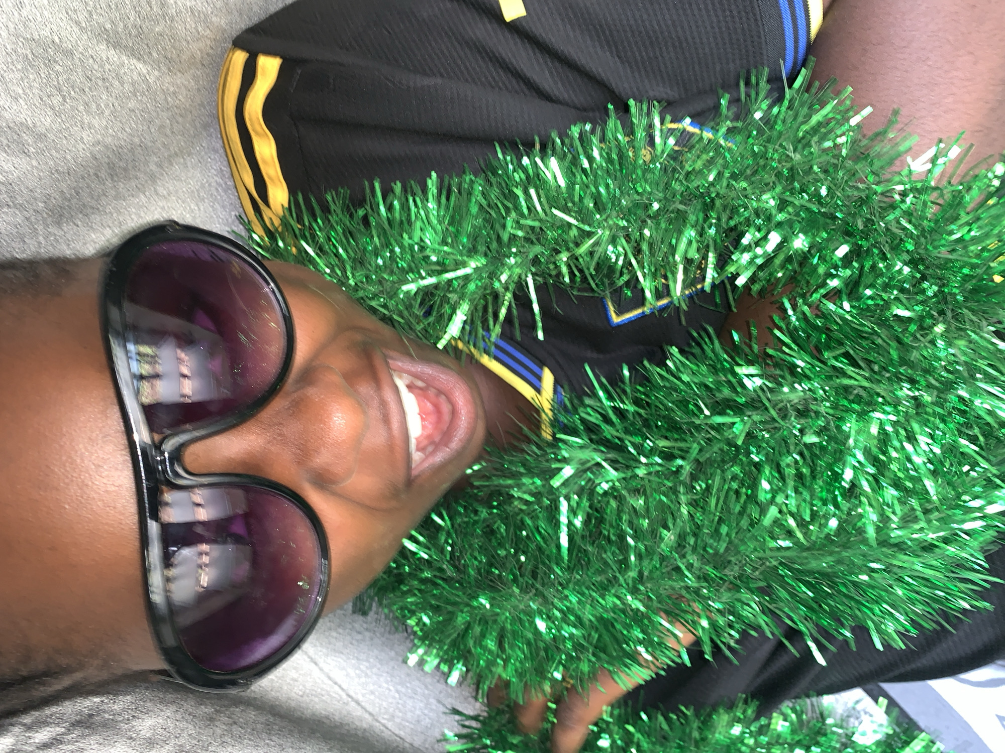 A young Black individual wears large, dark sunglasses and a green tinsel garland around their neck. They are smiling broadly, showing their teeth, and appear to be looking at the camera. The individual is wearing a black t-shirt with yellow and blue stripes on the sleeves. The background is a textured gray surface, possibly a wall or fabric. The image does not provide any specific information about the location in the city of Rafin Par, Nigeria.