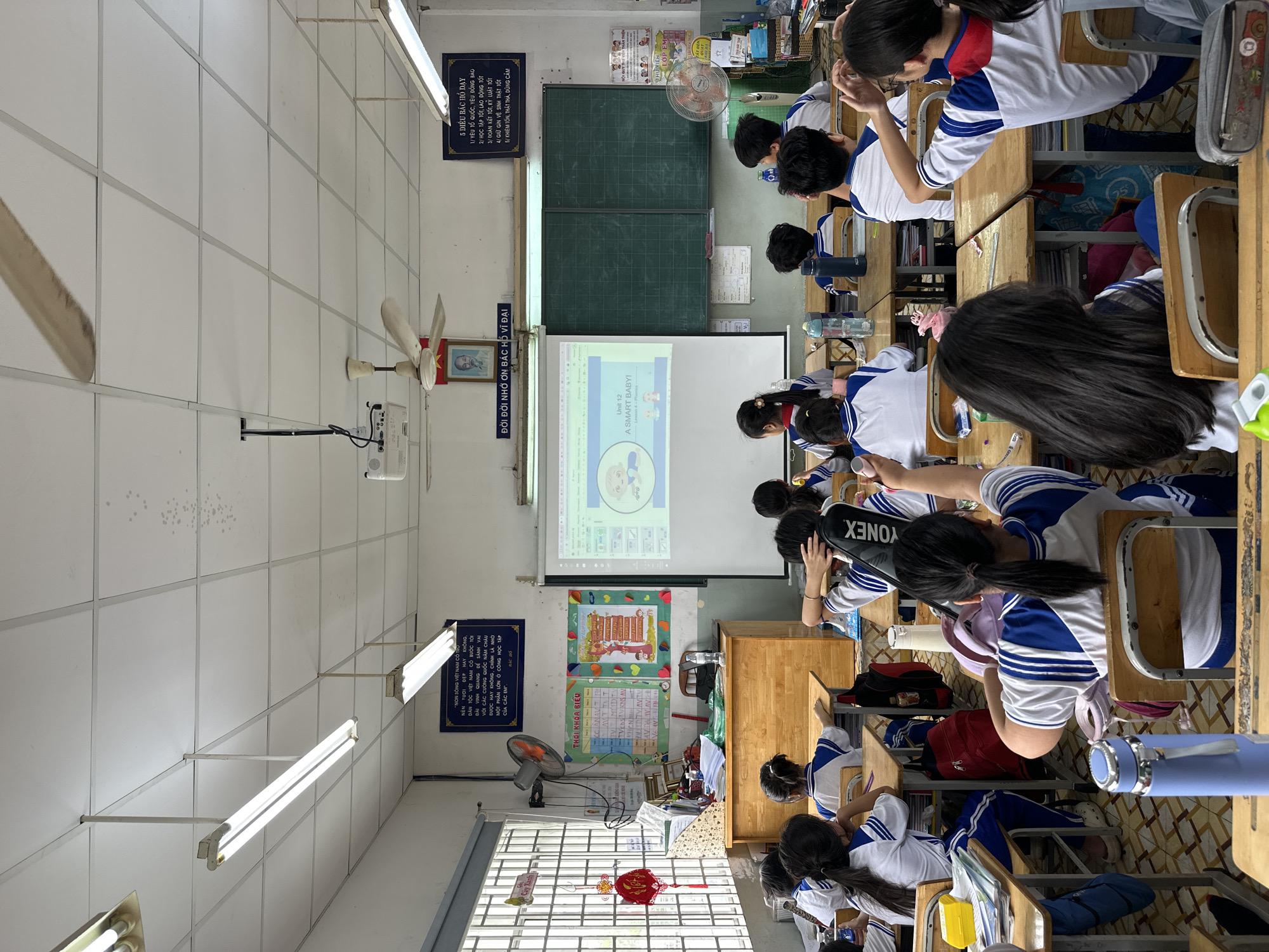 The image shows a classroom in Ho Chi Minh City, Vietnam, where a teacher is giving a lesson. Students wearing school uniforms are seated at desks facing the projector screen where the lesson is displayed. Classroom decorations include a chalkboard, fans, posters, and an image of Ho Chi Minh. A sign outlining "5 Điều Bác Hồ Dạy" (5 Things Uncle Ho Taught) is visible on the wall. The classroom appears to be in session as students are looking towards the screen.