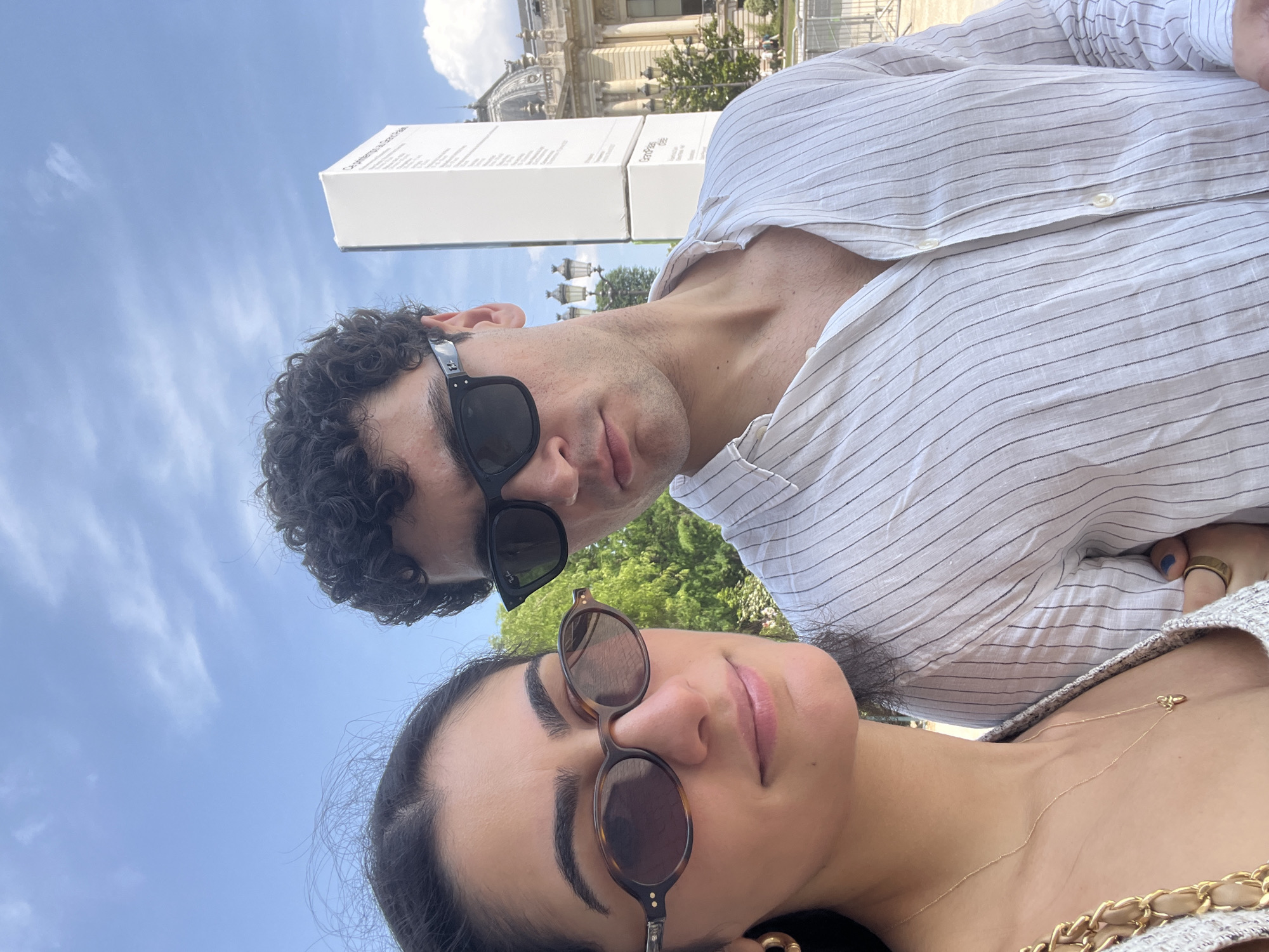A young couple is posing for a selfie outdoors in Paris, France. The man, on the right, is wearing a white pinstripe shirt and black sunglasses. The woman, on the left, is wearing a grey top and tortoise-shell sunglasses, and a gold necklace with a small pendant. In the background, a tall white sign stands in front of a building with a domed roof. The sky is blue with scattered white clouds, and trees are visible to the left and right. This appears to be the Grand Palais in Paris.