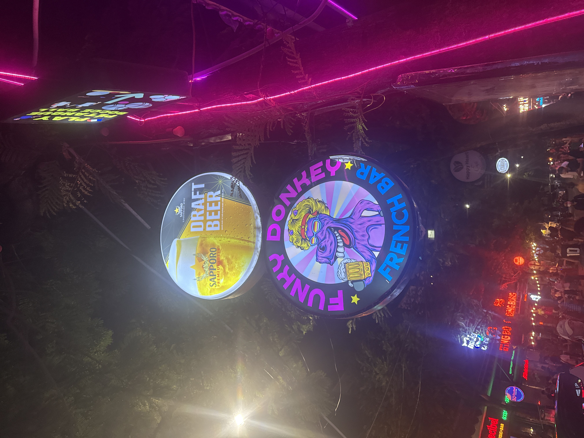 Here is a matter-of-fact description of the image:
This appears to be an outdoor commercial scene at night, potentially in a tourist area. The dominant features are two illuminated signs for bars/restaurants. One sign, circular in shape, is for "FUNKY DONKEY BAR FRENCH," featuring a cartoonish donkey character. The other sign promotes "DRAFT BEER," with "SAPPORO PREMIUM BEER" prominently displayed.
In the background, there is a dense crowd of people, suggesting a busy area. Several other illuminated signs and establishments are visible, including "BINGBOEING BONG" and "Decibel MUSIC+BAR GOODS". The image also includes bright pink neon lights above the signs and what looks like fern plants. The place is well lit up with lots of different signs and lights.