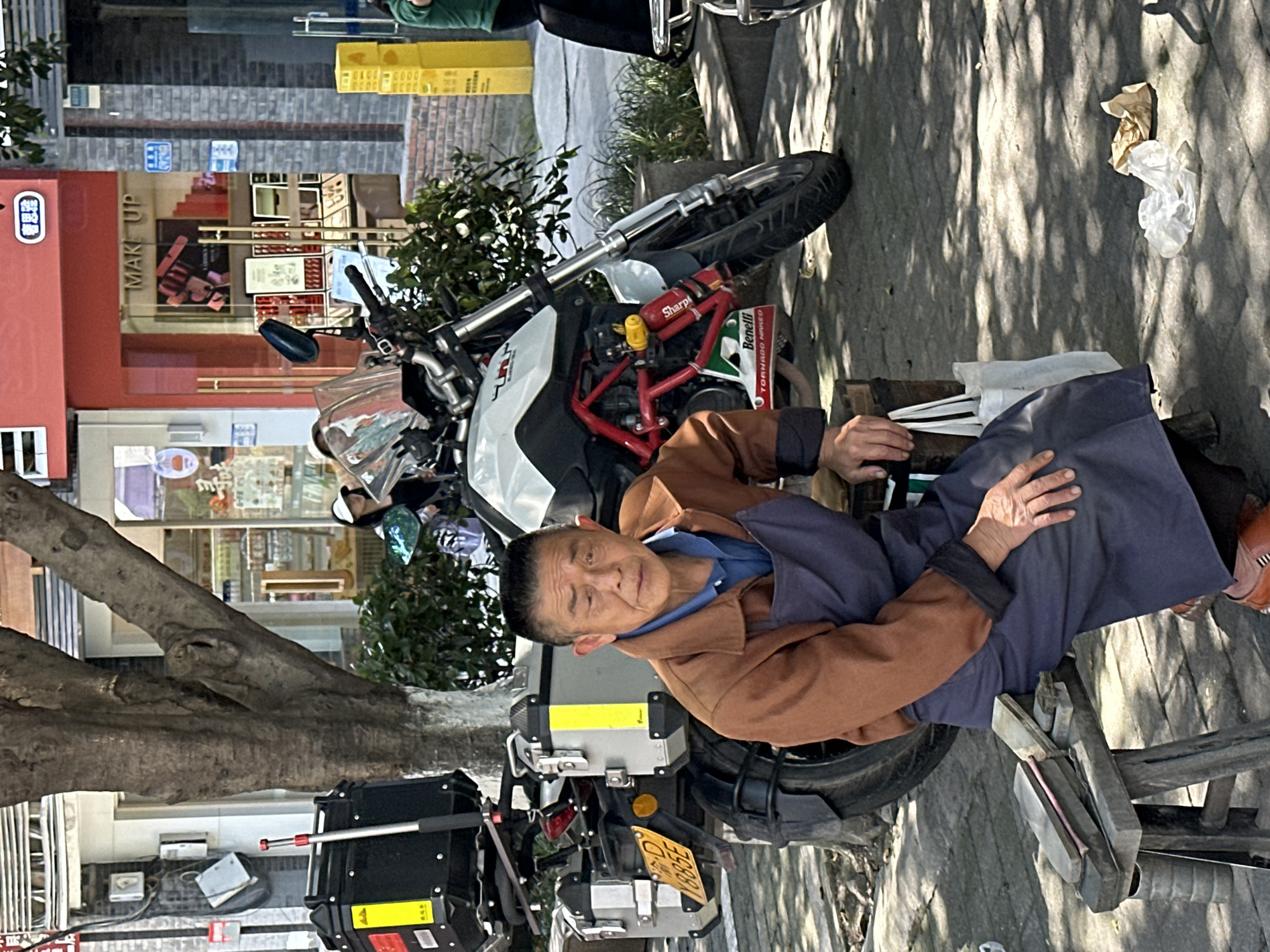 An elderly man sits on a weathered wooden bench beside a motorcycle in the Shapingba District, China. The man wears a brown jacket over a blue shirt and dark pants. His hands rest on his lap. The motorcycle has white and red details, with a license plate partially visible, reading "渝 885E." A black rectangular container is attached to the back of the motorcycle. Behind the motorcycle is a tree, and in the background, buildings are visible, including one with the English words "MAKE UP." The scene is bathed in sunlight, casting shadows on the pavement.