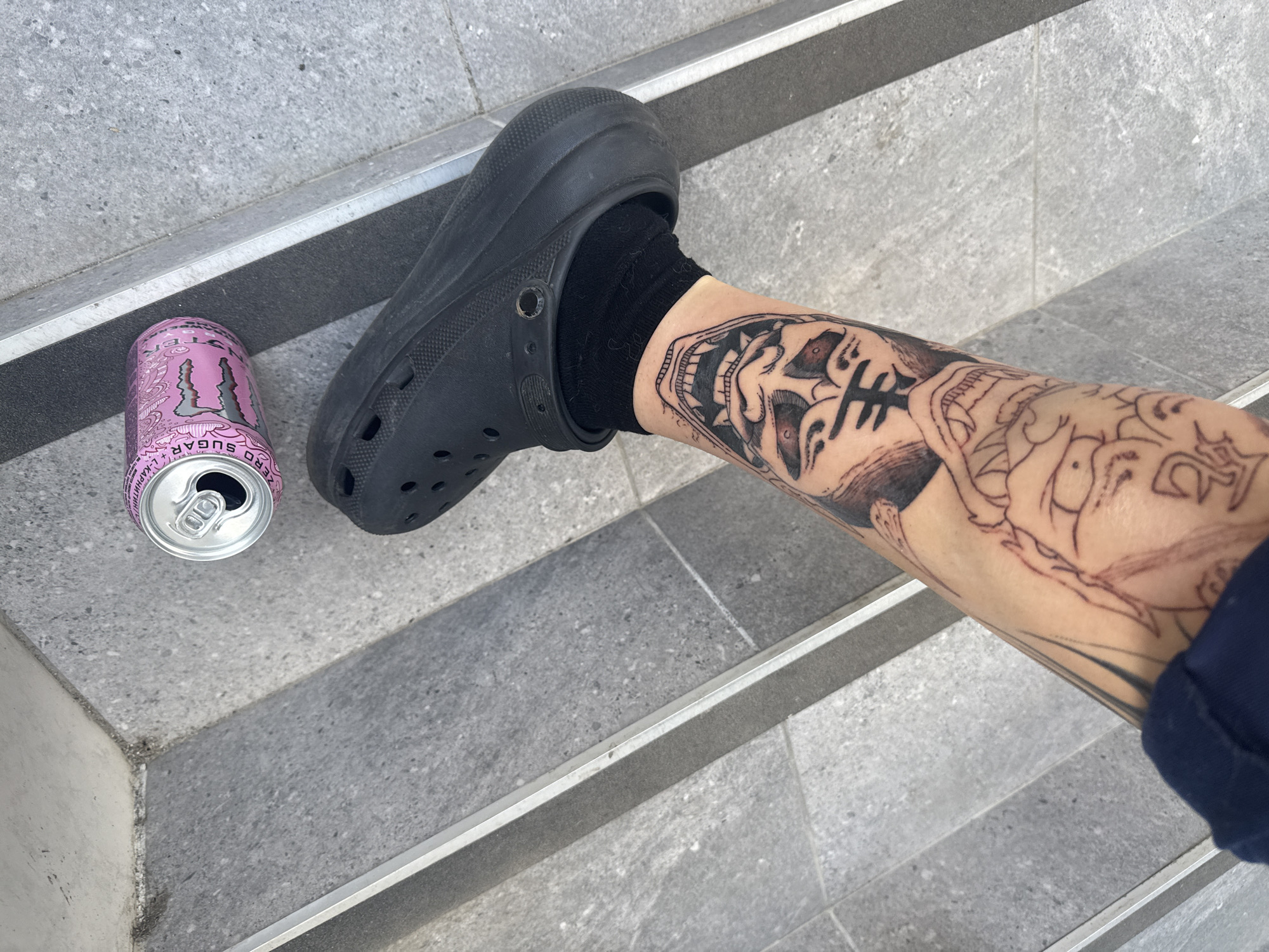 On a grey set of steps, a person's leg is seen wearing a black sock and a black Croc-style shoe. The leg has multiple tattoos. An empty pink Monster energy drink can sits near the shoe. The location is unknown and is not discernable from the image.