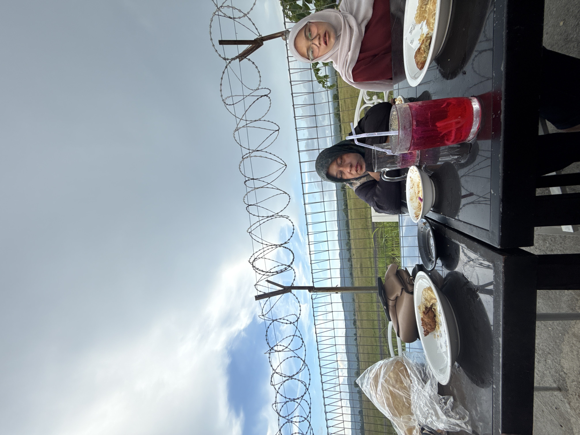Two women are seated at a black table outdoors in Cotmalem, Indonesia. The woman on the left wears a dark green hijab and a black jacket. She is looking downwards. The woman on the right wears a light pink hijab and a maroon shirt. She is wearing glasses and looking slightly to her left. In front of them are plates with food, drinks, and cutlery. A glass of red liquid with straws is positioned in the center of the table. A bag and a plastic-wrapped loaf of bread are on the left side of the table. Behind the table is a fence with barbed wire. The background shows a field and hills under a cloudy sky.