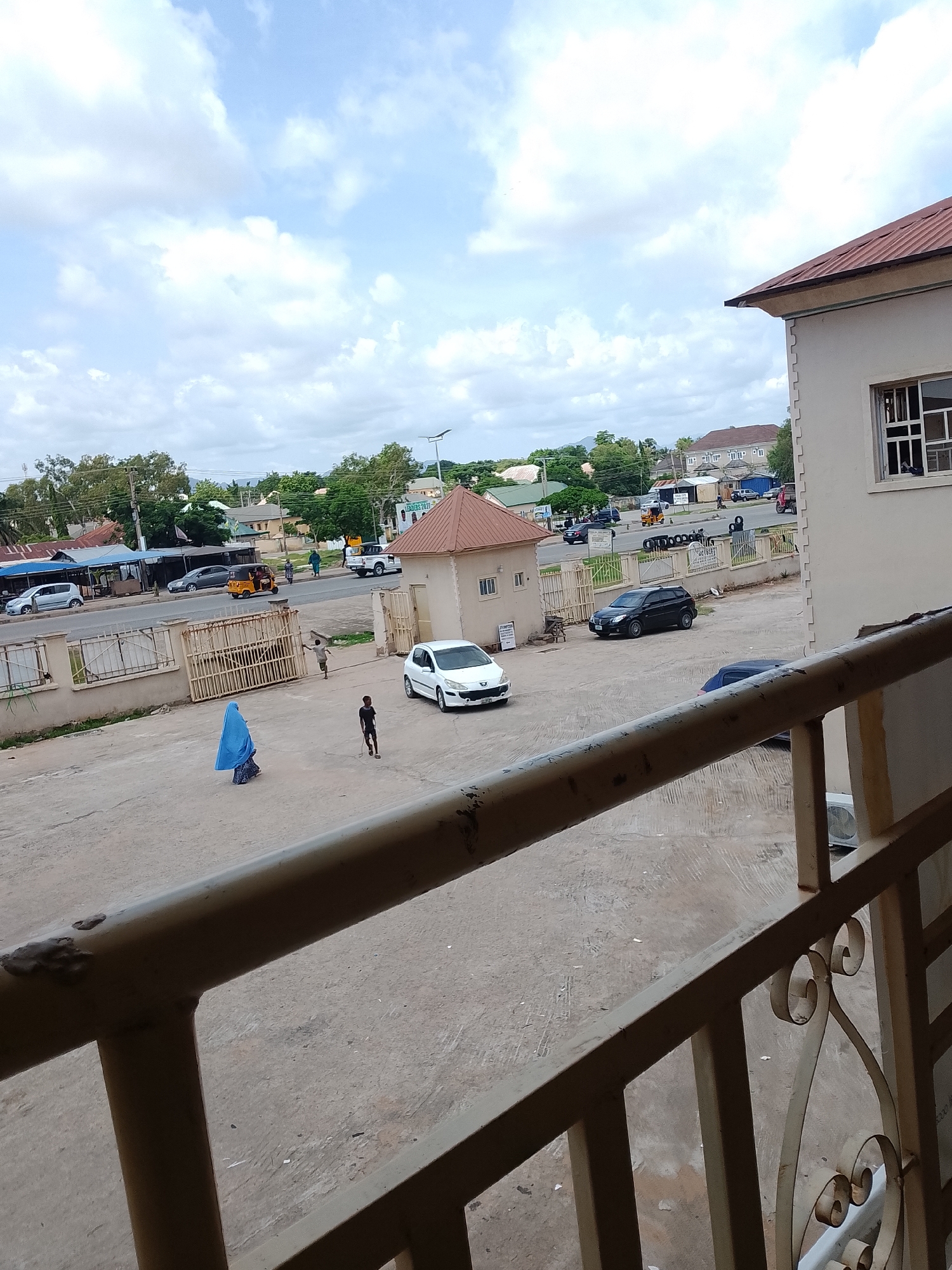 The image captures an outdoor scene in Jalingo, Nigeria, with a building in the foreground and a street with vehicles and people in the background. A light-colored residential building with a reddish-brown roof occupies the right side of the frame. Its exterior wall is textured and features a window with multiple panes. Below the building, a paved area serves as a pathway or courtyard, with a railing in the immediate foreground.
The middle ground shows a paved street with various vehicles, including a yellow auto-rickshaw, a white SUV, and a dark-colored car. Several people are visible on the street and on the paved area. One person, wearing a blue hijab and patterned skirt, walks across the paved area. Another person, a child in a black t-shirt and shorts, is seen holding a stick. Further down the street, more individuals are walking and interacting. The background reveals more buildings, trees, and a cloudy sky. The overall environment suggests a typical urban setting in Nigeria, likely during daytime.