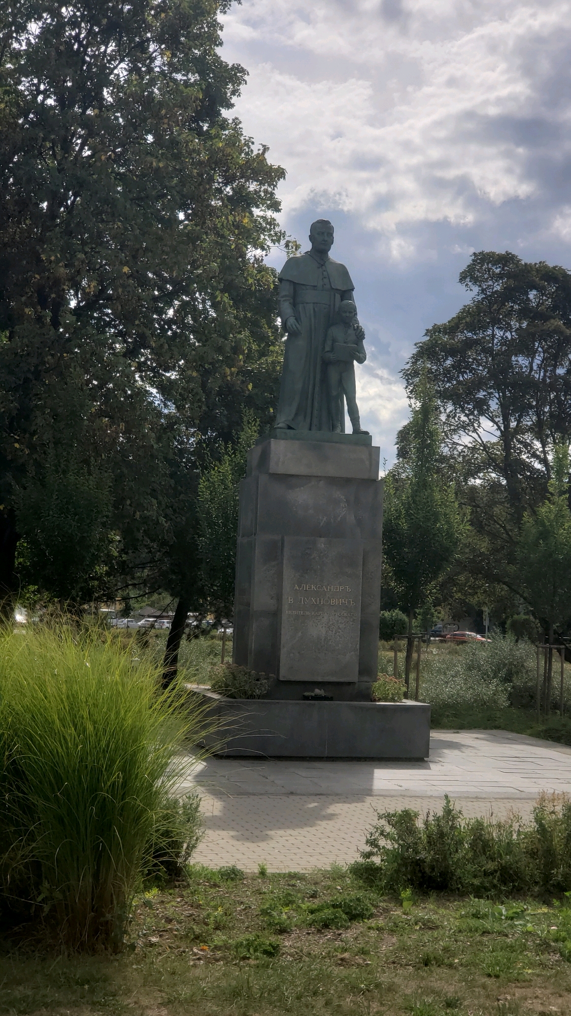 The image depicts a statue of a man in religious attire, possibly a priest, standing next to a young boy. The statue is made of dark metal and stands on a large, multi-tiered stone pedestal. Inscriptions are visible on the pedestal, written in Cyrillic script. The statue is situated in an outdoor environment with lush green trees and grass surrounding it. In the background, there are more trees and a cloudy sky. The ground in front of the pedestal is paved with light-colored tiles. The overall scene appears to be a monument or memorial located in a public space, likely a park or square. This monument is located in the city of Prešov, Slovakia, and it commemorates Alexander Duchnovič, a writer and priest.