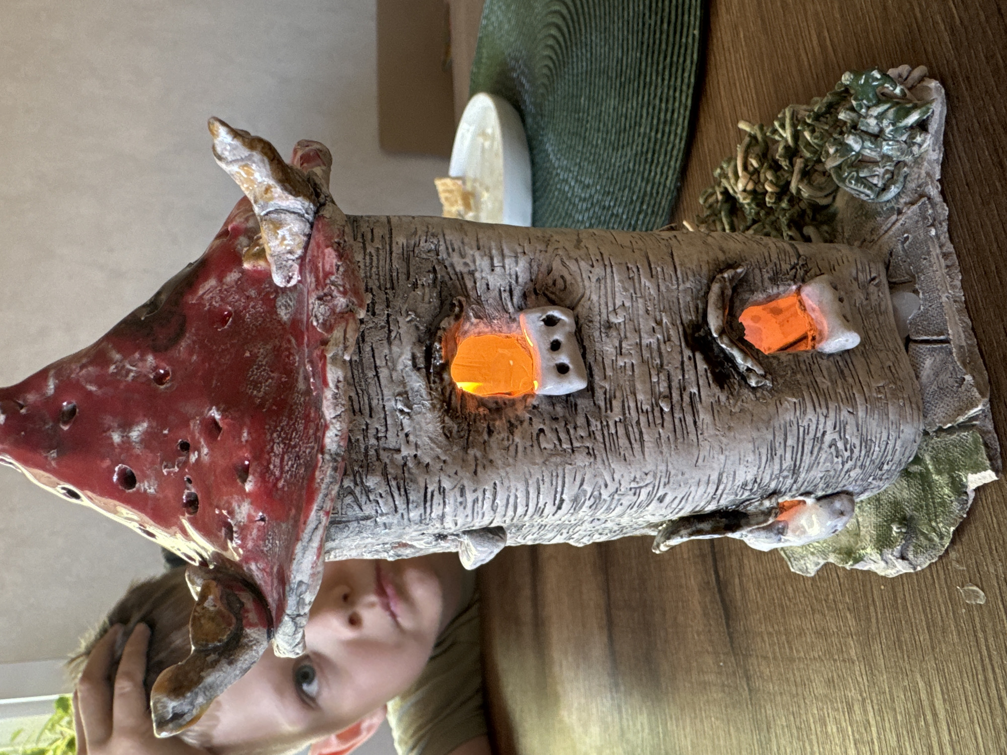 A ceramic house is centered in the frame, featuring a red, pointed roof with holes and a textured gray body that resembles bark. Light emanates from small window openings. At the base, textured green ceramic elements mimic foliage. A blurred image of a child’s face is visible beneath the house, likely reflected from a surface. The house sits on a brown, wood-grained surface, possibly a table. Additional household items are partially visible in the background, including a plate and a green placemat. The location is Gornyi (Dvurechenskoe S/P), Russia.