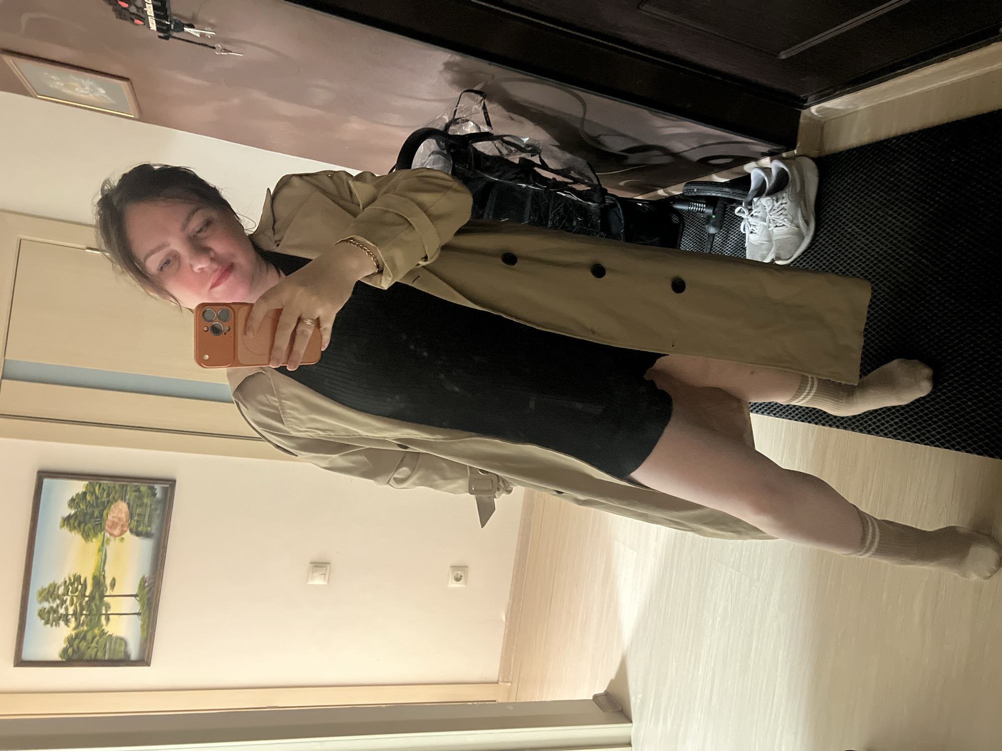 A woman is taking a selfie in what appears to be an apartment or house interior. She is wearing a black dress, a beige trench coat, and striped socks. Her right hand is holding an orange phone.
In the background, there's a hallway, a door, a stroller with a rain cover, and a pair of grey sneakers. On the walls are a picture in a frame and electrical outlets. The location is likely a residential building in Perm, Russia, judging by the interior design and style, although this is difficult to ascertain with full certainty without further evidence.