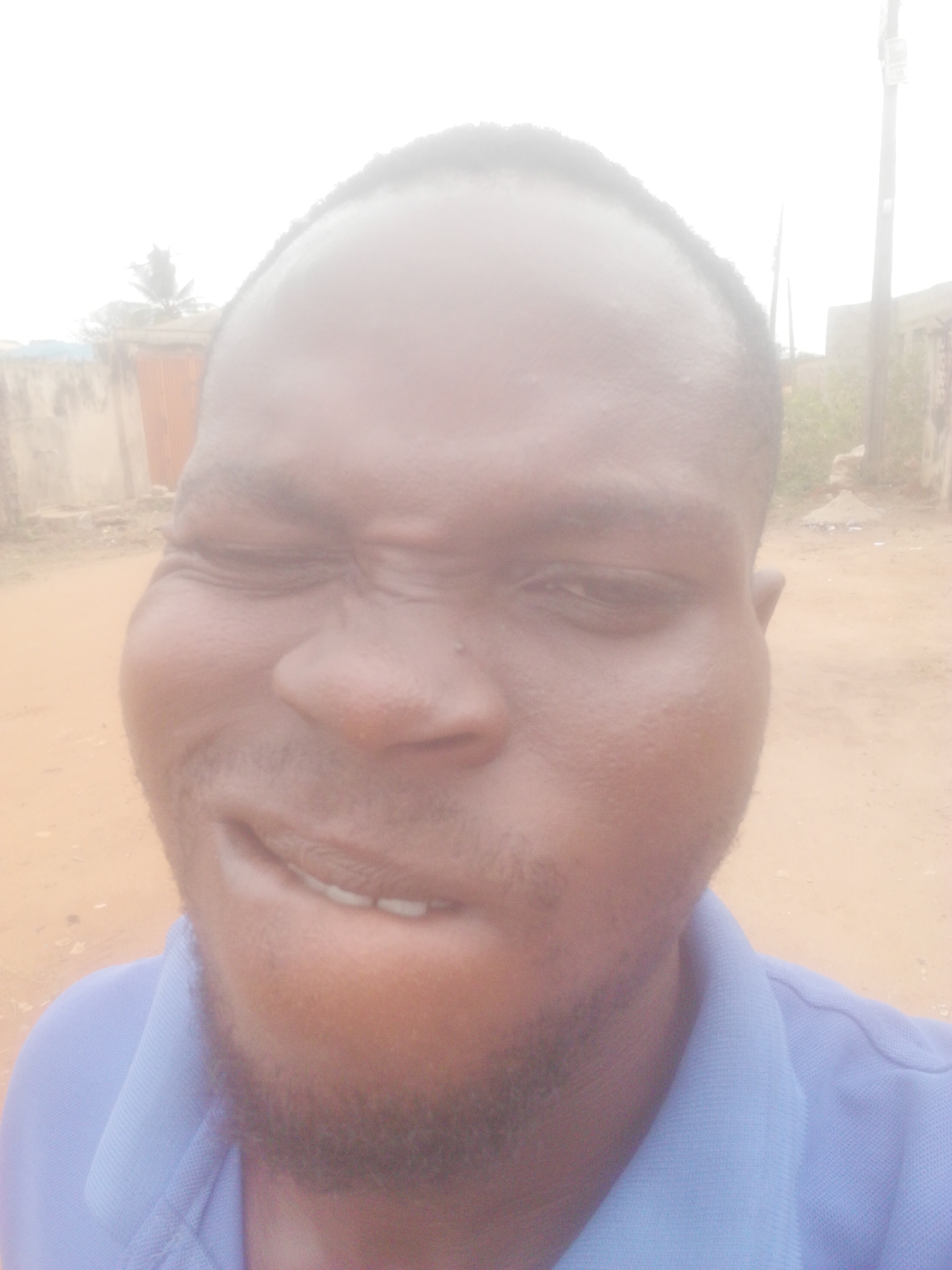 A man with dark skin and a beard is seen in a close-up selfie-style shot. His facial expression appears strained. He is wearing a blue polo shirt. The background suggests an outdoor environment in Ikorodu, Nigeria, with a dirt road and buildings visible in the distance. A telephone pole can be seen far away. The sky is overcast.
