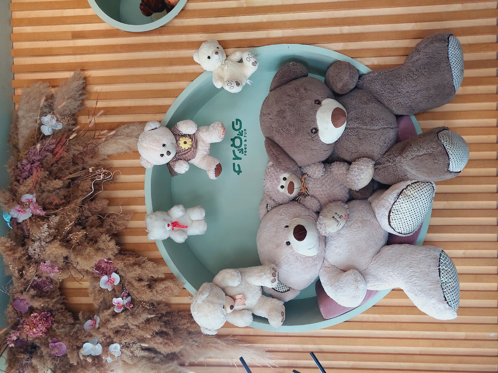 A collection of teddy bears are displayed on a teal shelf against a wooden paneled wall decorated with dried floral arrangements. The teddy bears vary in size and color, with some white, beige, and brown bears. One large beige bear is seated on the shelf, while a large brown bear sits next to it. Other smaller bears are positioned on the shelf and on the wall above it. The shelf has a logo that reads "FRÖG FOOD & FUN". This scene is located in Sofia, Bulgaria.