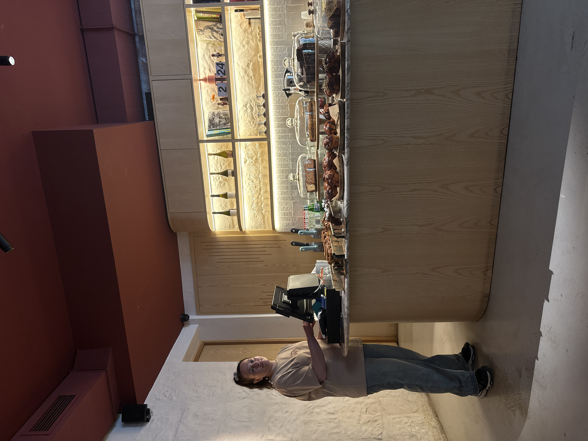 A person is standing behind a counter inside a cafe or bakery, possibly in Yerevan, Armenia, although the specific location is not definitively discernible from the image alone. The person is operating a point-of-sale system. The counter displays an array of pastries and baked goods under glass domes. Behind the counter is a lighted display of bottles, jars, and other items. The cafe features a modern design with red accent walls. The floor is light colored concrete or a similar material.