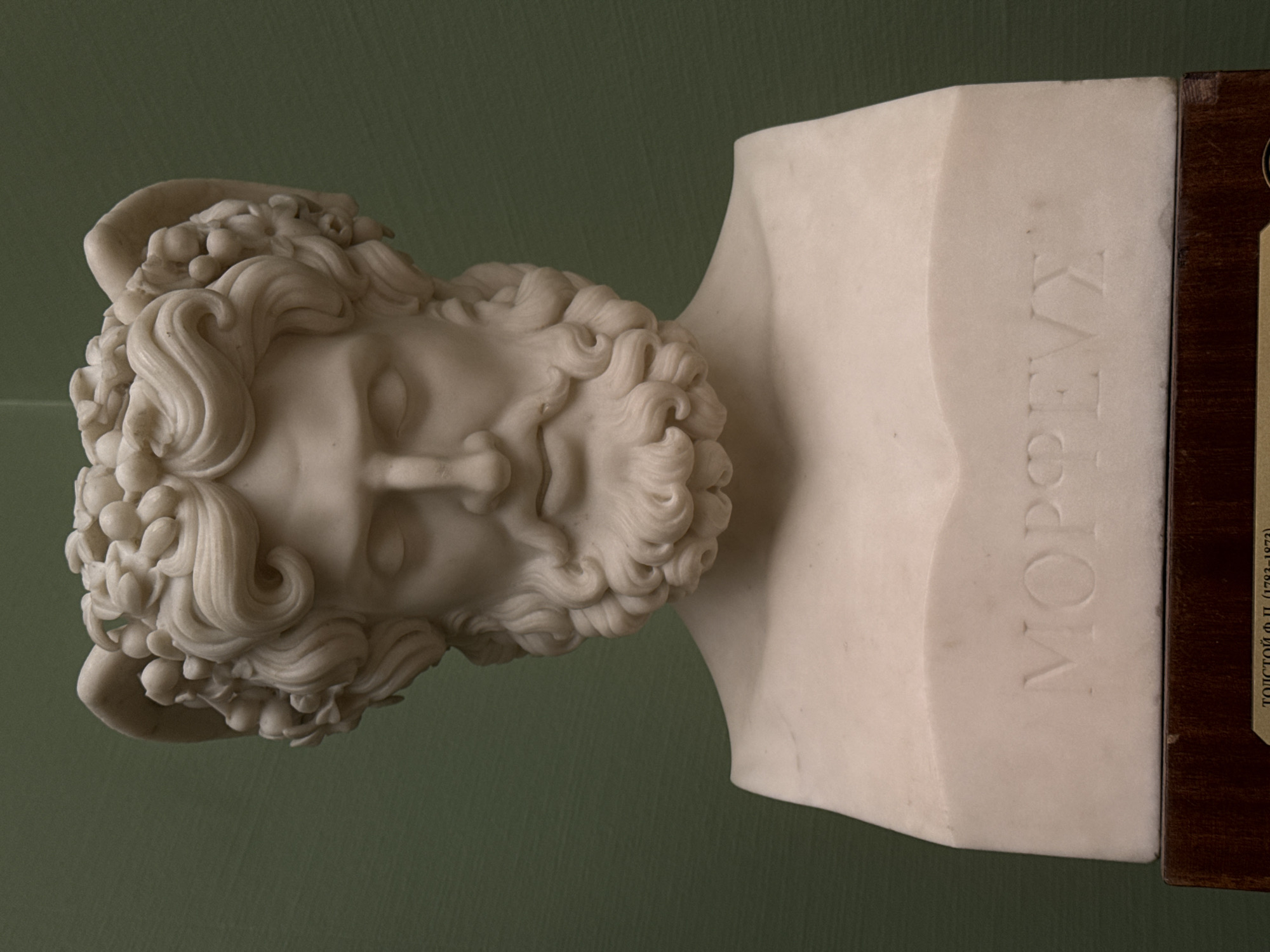 The image depicts a white marble bust of a bearded male figure with curled hair, possibly of classical or neoclassical style, bearing ram's horns. The bust is oriented on its side, revealing the name "ΜΟΡΦΕΥΣ" inscribed in Greek letters on the side of the plinth. A small portion of a wooden information plaque is visible to the right. This bust is located in the Pushkin State Museum of Fine Arts in Moscow, Russia.