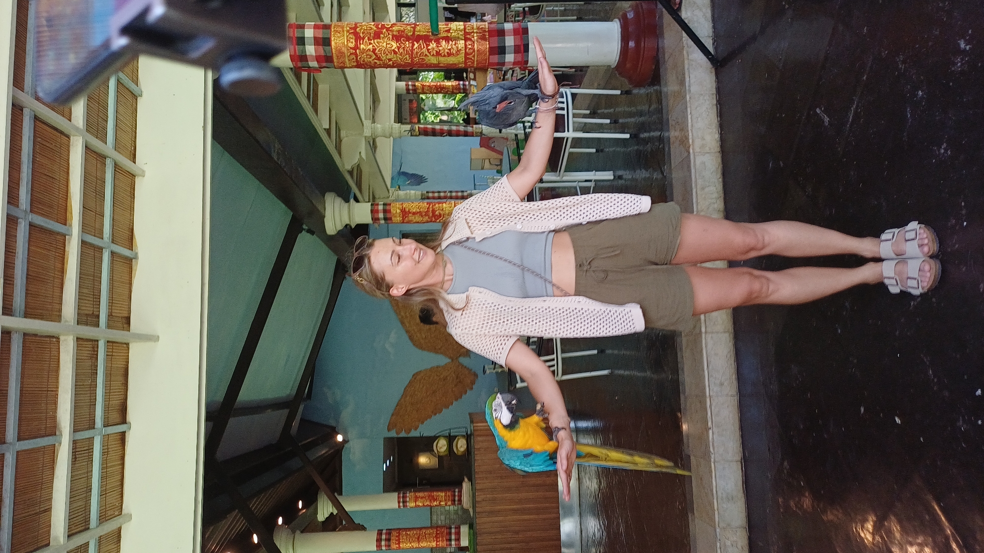 A full-length photograph shows a woman standing in what appears to be a bird park in Sukawati, Indonesia. The woman is light-skinned and wearing a light gray t-shirt, khaki shorts, a beige crocheted cardigan, and light-colored sandals. Sunglasses are pushed atop her head. She is standing with her arms outstretched. On her right arm perches a blue and yellow macaw. On her left arm is a gray bird, possibly a pigeon.
The background indicates an indoor setting. A mural of a bird with outstretched wings is visible behind the woman. To her right are white columns decorated with a red and gold patterned fabric. The floor is dark and reflective.
