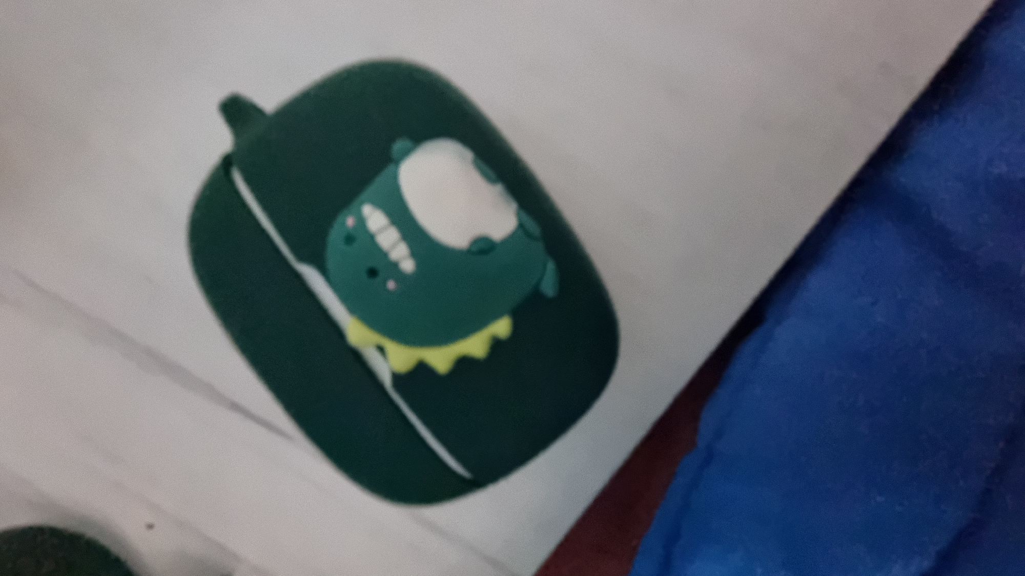 A close-up, slightly out-of-focus, and slightly angled shot shows a dark green case with a cartoon dinosaur figure on a light surface. The case appears to be for an electronic device, possibly earbuds, featuring a small loop at the top. The cartoon dinosaur is primarily dark green with a lighter, cream-colored belly and a yellow frill. Part of a blue item (possibly clothing or a blanket) is visible in the lower right corner. The location is undetermined.