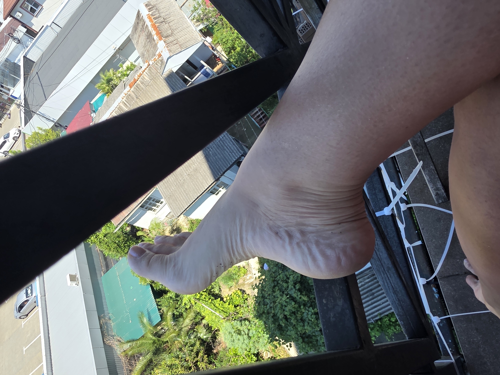 The image shows a close-up of a person's foot and lower leg, positioned on a dark metal railing overlooking a city. The background reveals a dense urban environment with various buildings, rooftops, and lush green vegetation, suggesting a warm climate. Some buildings have corrugated metal roofs, while others are lighter in color. A silver car is visible parked in a lot in the upper left portion of the image. Zip ties are attached to the railing in the foreground. The lighting indicates it is daytime. The specific location of Sochi, Russia, is not definitively identifiable from the visual elements alone, as the scene depicts a generic urban landscape with typical features found in many cities.