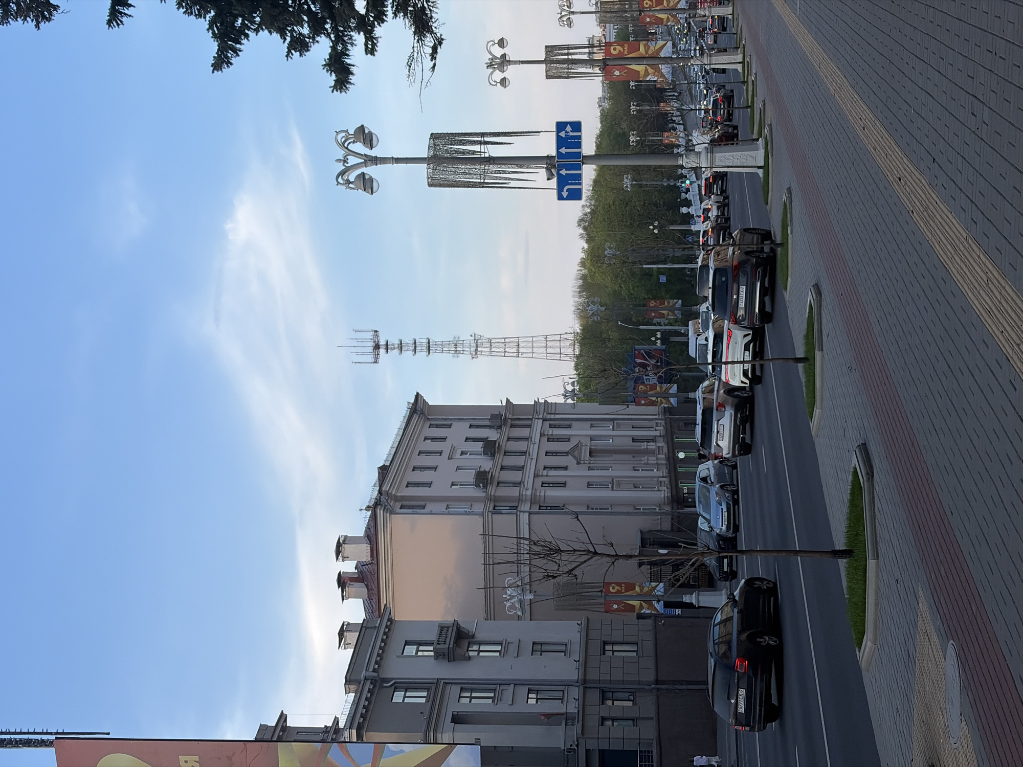 The image shows an urban street scene in Unknown, Unknown. A multi-lane road is lined with parked cars on one side. Buildings of varying heights line the other side of the street. Streetlights with decorative elements are present along the road. A tall metal lattice tower is visible in the distance. The sky is blue with some clouds. There are various signs and banners visible. The sidewalk is paved. There are no visible people in the image.