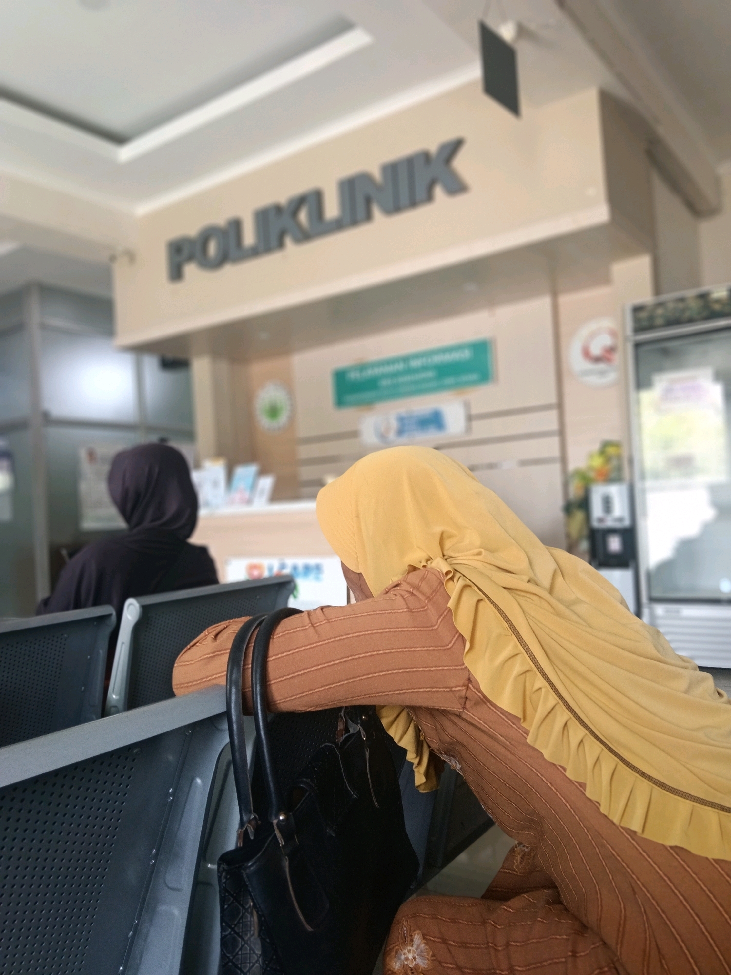 The image shows a waiting area at a Poliklinik in Balun, Indonesia. In the foreground, two individuals are seated, facing away from the camera. The person on the right wears a yellow headscarf and a brown garment with pinstripes and ruffles. A black handbag with a quilted texture hangs from their arm. The person on the left wears a black headscarf and garment. They are seated on grey chairs arranged in rows. In the background, a sign with "POLIKLINIK" is visible above a reception or service counter. Other signs and a water dispenser are also present in the background, along with glass partitions and a refrigerator. The overall environment appears to be an indoor waiting room, possibly a clinic or hospital.