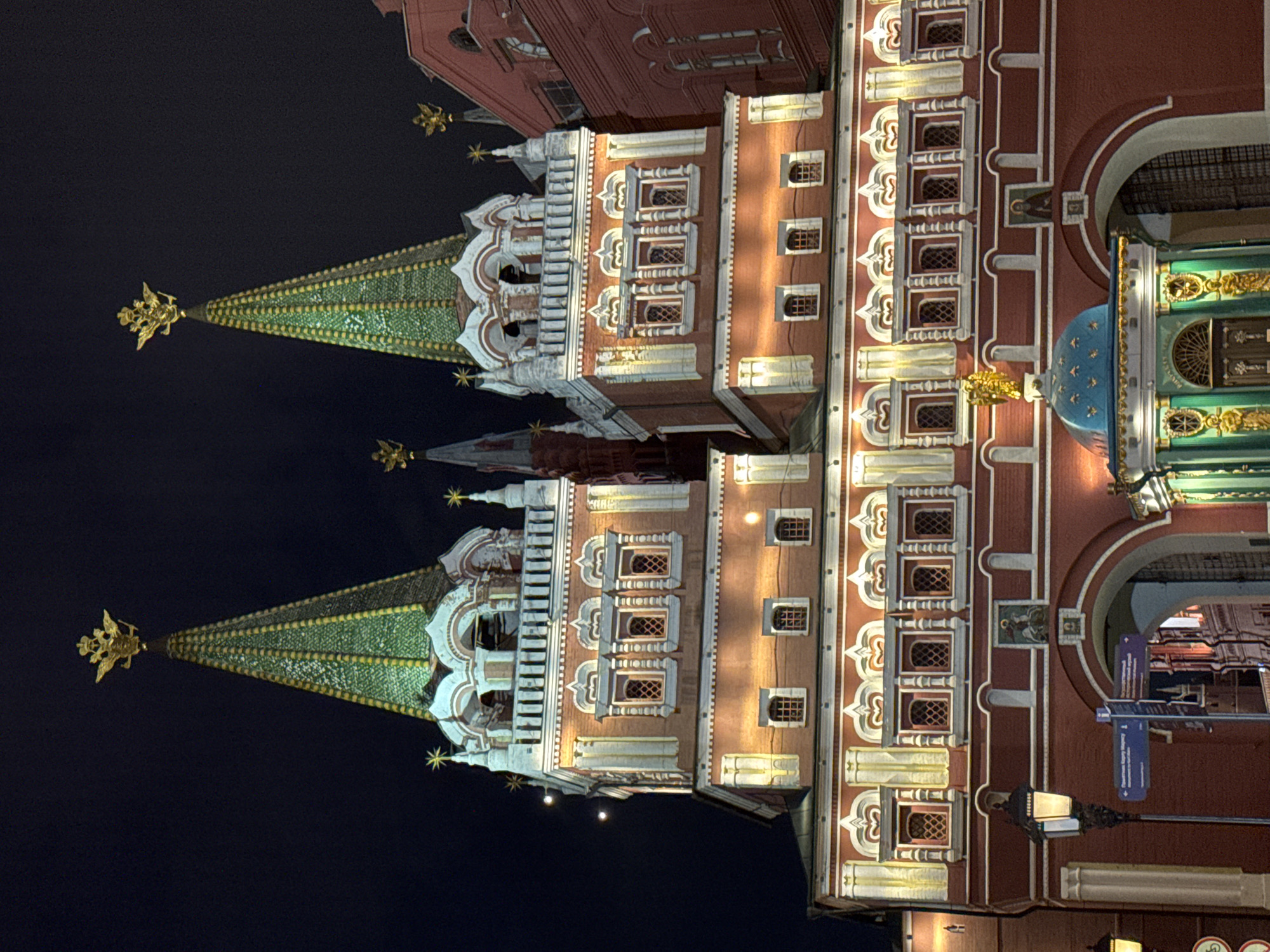 The image shows the Resurrection Gate in Moscow, Russia, at night. The gate is illuminated and its red brick structure with white detailing is prominent. Two towers with green spires topped by golden double-headed eagles dominate the skyline. Below the towers, there are multiple levels of windows and decorative architectural elements. In the foreground, a small, ornate blue-domed structure with golden figures is visible. A lamppost stands to the left of the gate. The sky is dark, indicating nighttime.