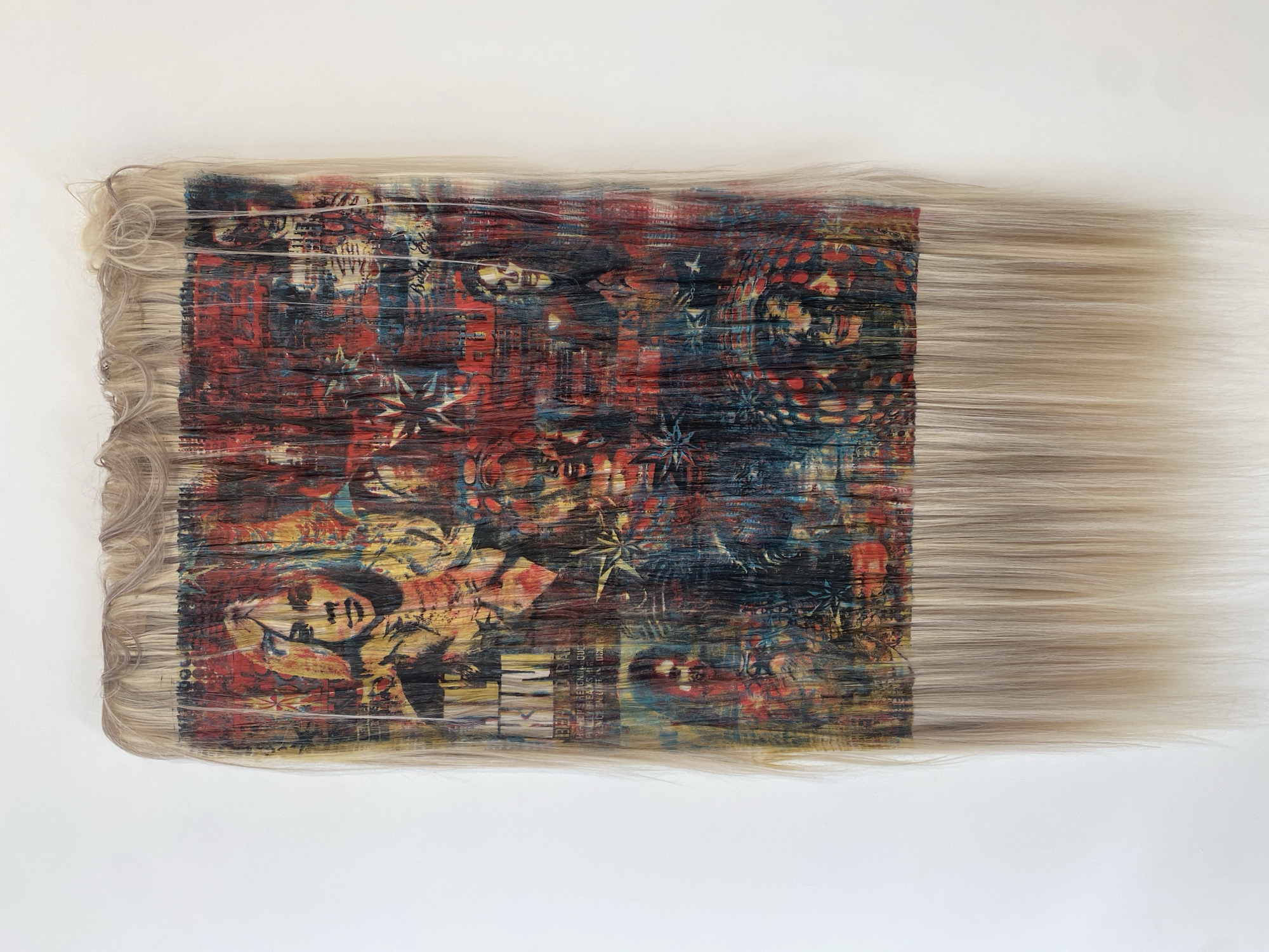 The image shows a piece of abstract art. The artwork has a square core filled with a complex design featuring red, blue, yellow, and black colors. Within the design are recognizable shapes that resemble faces, stars, and other indistinct forms. Encircling the square core are long, light-blonde strands of hair, creating a frame around the design. The hair extends outward, giving the impression of movement or flow. The artwork appears to be hanging on a plain white wall. There is nothing in the image to suggest its location is in Paris, France.