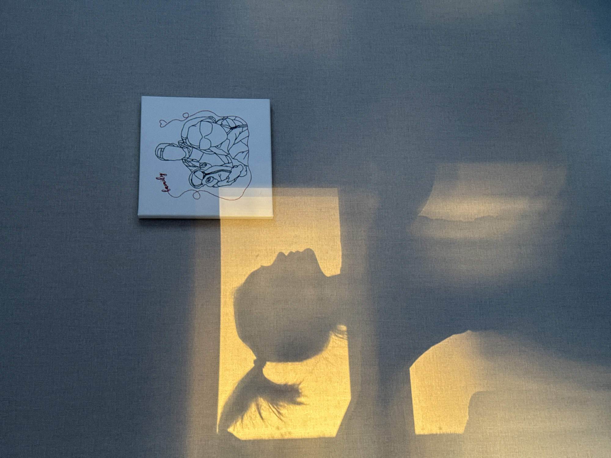 The image captures a serene indoor scene in Izhevsk, Russia, characterized by warm, natural light and subtle details. The primary subjects are a small, square white canvas hanging on a textured grayish-blue wall, and the distinct shadow of a person cast by sunlight.
The canvas features a minimalist black line-art drawing. It depicts two figures, possibly a couple, with intertwined shapes and small heart motifs. Below the drawing, in red cursive script, is the word "family." The canvas itself is slightly illuminated by sunlight from the bottom right.
Dominating the lower and right portions of the frame is a strong, golden rectangle of sunlight on the wall. Within this illuminated area, a clear silhouette of a person's profile is cast. The shadow shows the head and upper body of what appears to be a child or young person looking upwards, with hair tied back in a ponytail or bun, discernible by the wispy details at the base of the neck. Other, more abstract shadows and light patterns are visible on the wall, suggesting geometric shapes from a window or architectural elements.
The lighting indicates either early morning or late afternoon, as the sun's low angle creates long, dramatic shadows and a warm, golden hue. The overall mood is peaceful and artistic, highlighting the interplay of light, shadow, and simple artwork. The wall has a visible fabric-like texture.