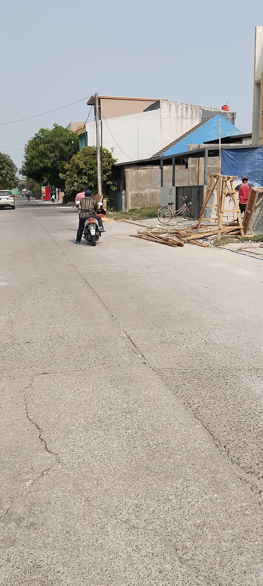 The image shows a street scene in Satriamekar, Indonesia, on a clear day. In the foreground, a grey asphalt road has visible cracks and a central seam. A person is riding a scooter with a passenger in the middle of the road, traveling away from the viewer. The scooter is white with a license plate. To the right of the road, there is a construction area with stacks of wood, a ladder, concrete blocks, and a blue tarp. A bicycle is parked near the construction materials, and a person in a red shirt is standing nearby. In the background, there are several buildings, including a light-colored building with a dark trim and a blue-roofed structure. Trees with green foliage are visible to the left, and a white car is parked on the left side of the street. The sky is light blue and clear.