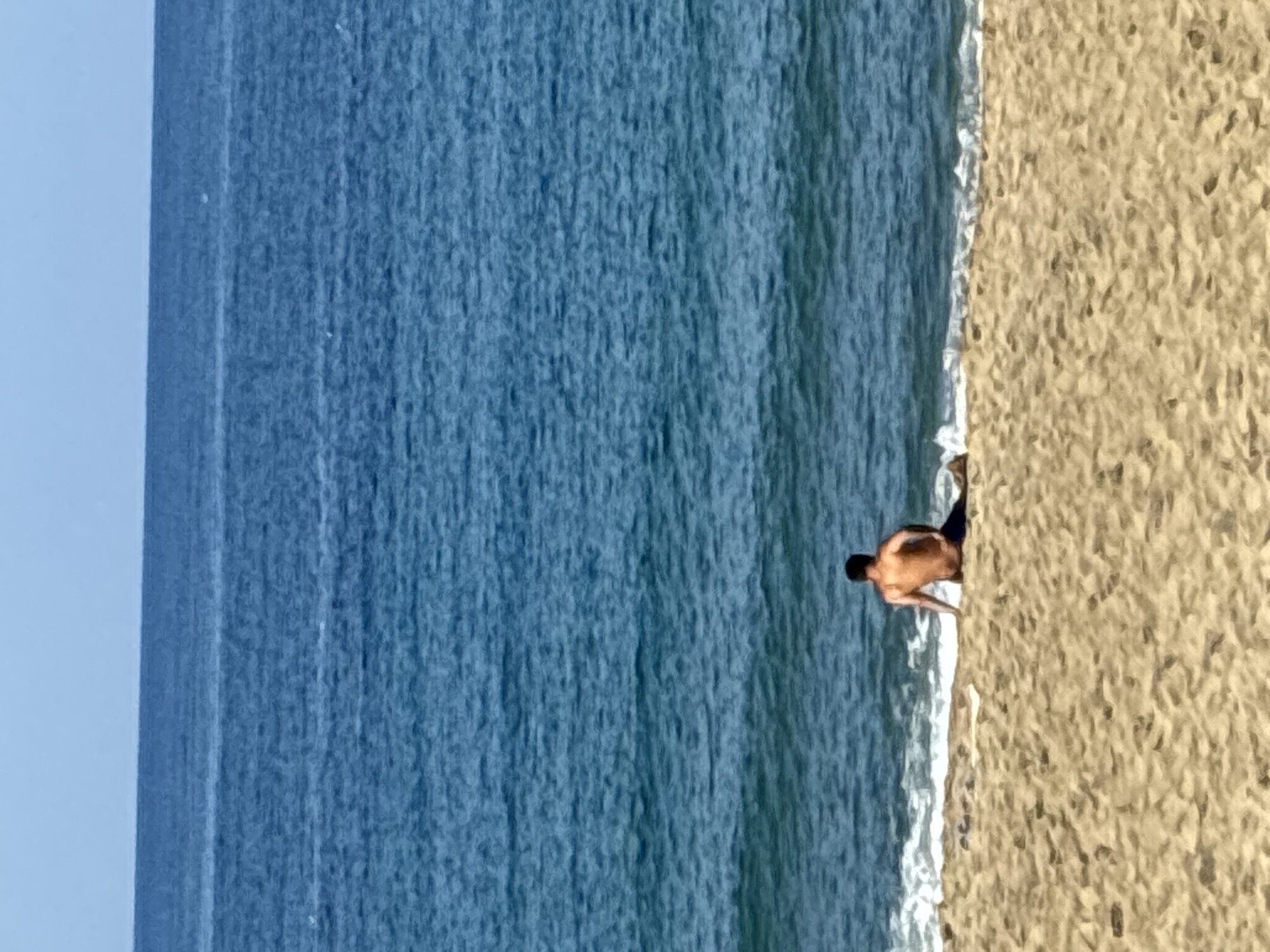 The image depicts a serene beach scene, likely captured in Alanya, Turkey, under clear daylight conditions. The composition is divided into three distinct areas: a light blue sky occupying the top portion, a wide expanse of deep blue ocean in the middle, and a stretch of golden-beige sand running along the right side. The ocean surface displays a textured pattern, indicative of gentle waves or ripples. A thin, white line of foamy surf marks the precise point where the water meets the shore.
In the lower right section of the frame, a single individual, appearing to be a man, is positioned at the water's edge. He is shirtless and wears dark swim shorts. His back is mostly visible to the viewer as he is either partially submerged, kneeling, or bending over. His posture suggests he is interacting with the shallow water or the sand, possibly wading, searching, or engaged in an activity near the surf. The bright lighting and clear sky indicate a sunny day. No text is visible in the image.
